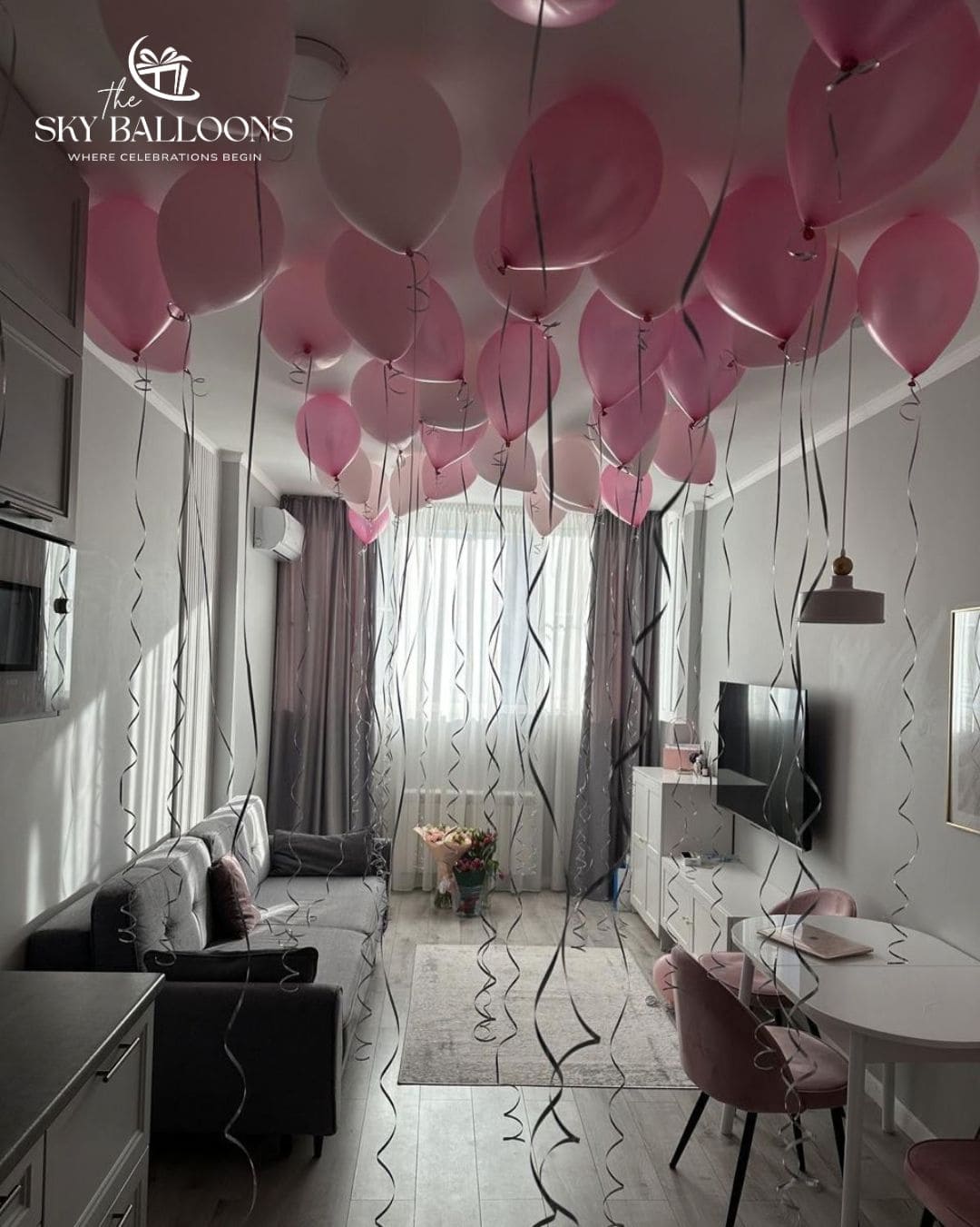 A decorated living room filled with numerous pink and white balloons floating near the ceiling, with long black confetti streamers hanging down. The room contains a gray sofa, a white dining table with pink chairs, gray curtains, and a TV on a white cabinet.