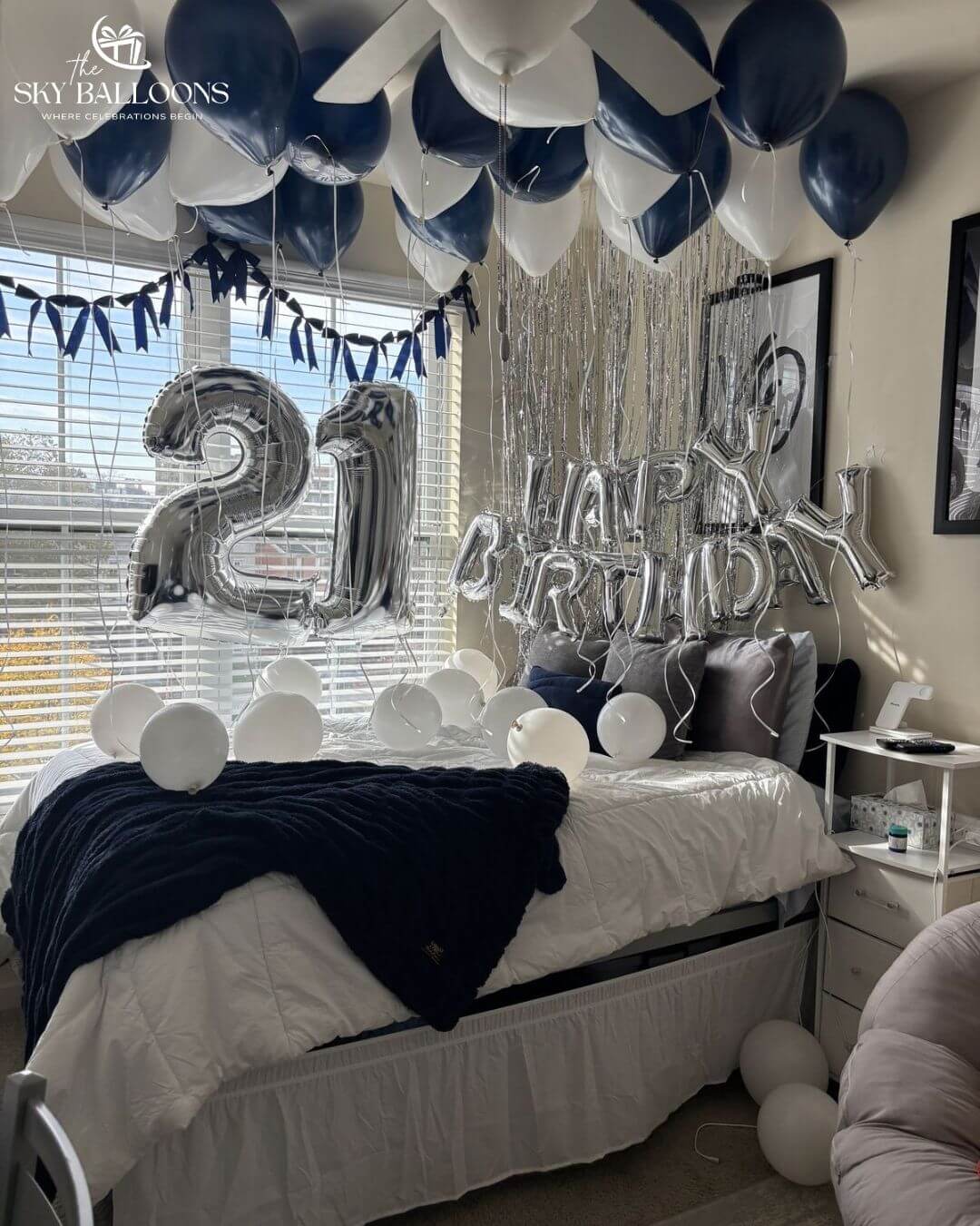 A decorated bedroom with a 21st birthday celebration setup, featuring silver and navy blue balloons, silver tinsel streamers, a large silver '21' balloon, 'HAPPY BIRTHDAY' letter balloons, and a navy blanket on a bed.