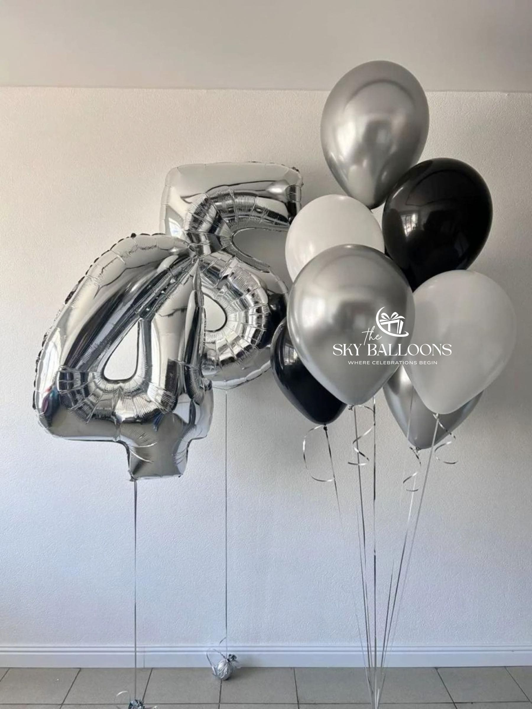 A cluster of metallic and matte balloons, including a large foil number 4 balloon and a bunch of round balloons in silver, black, white, and light gray, tied to a stand against a plain indoor wall.