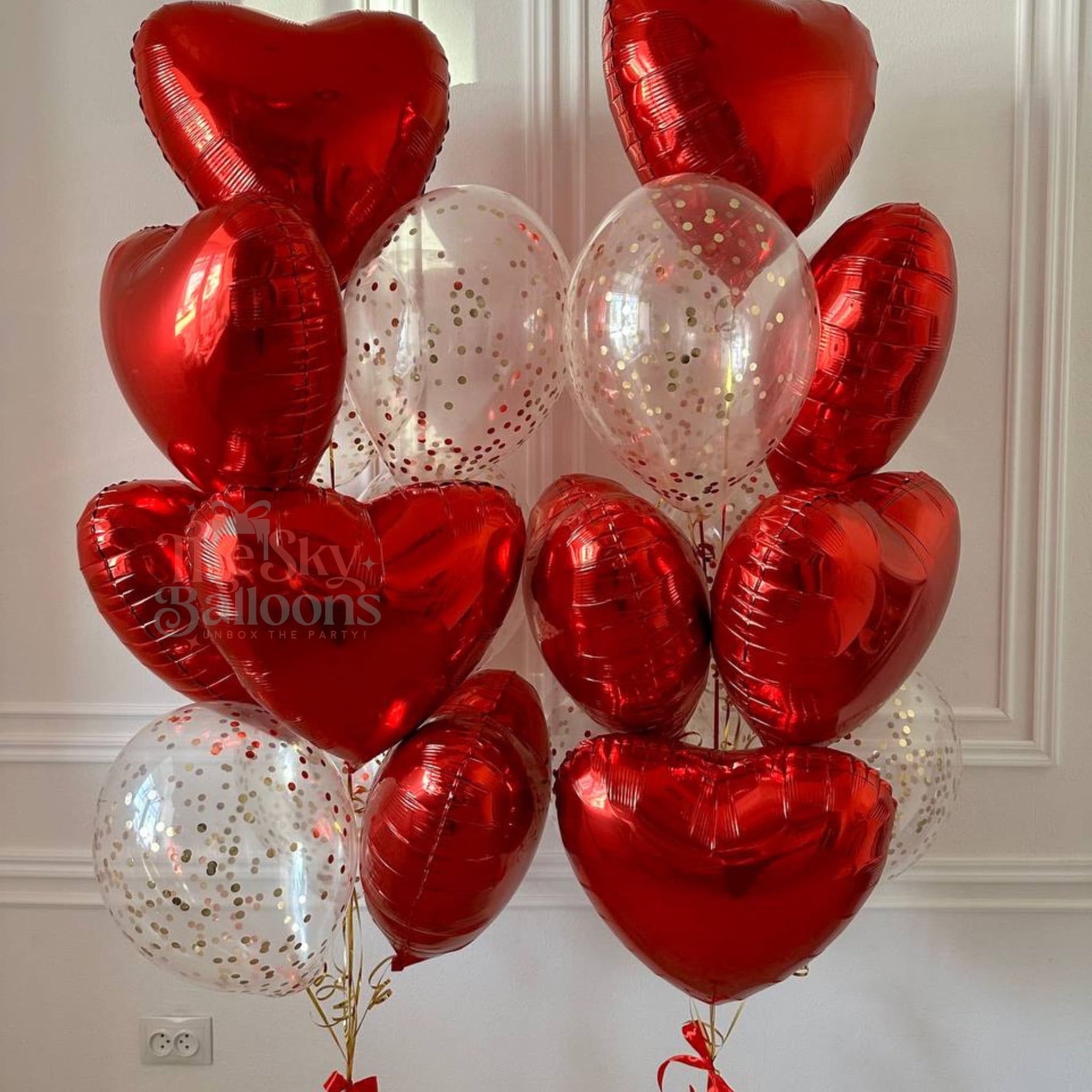 Bouquet of red heart-shaped balloons with clear balloons featuring gold confetti against a white wall.