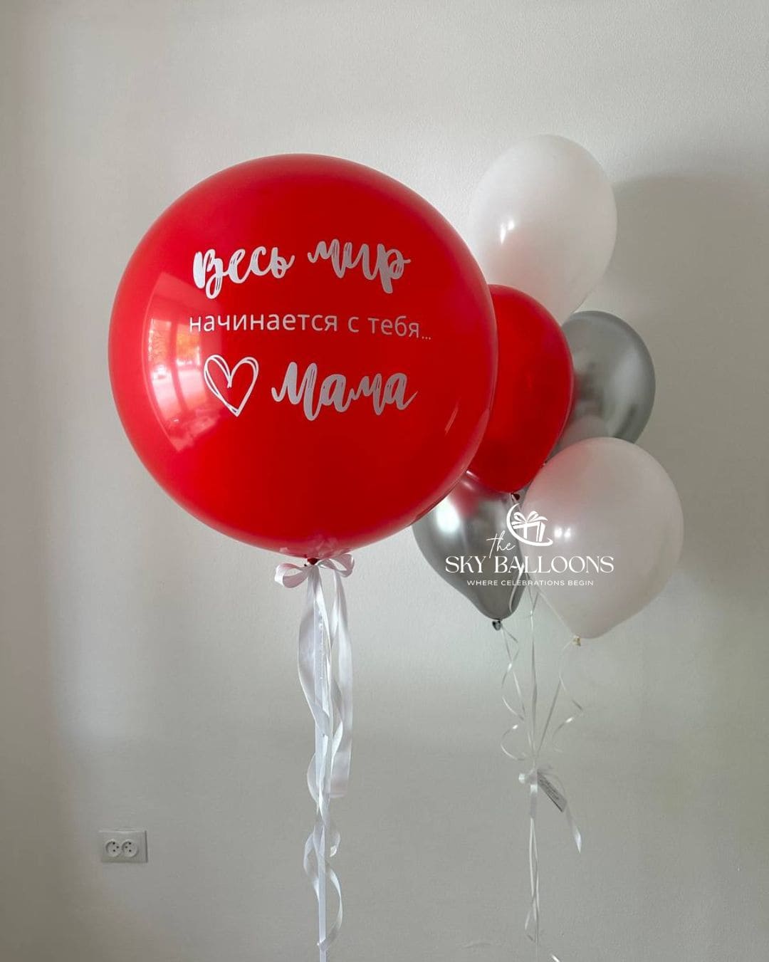 Personalised jumbo red balloon with a white Russian message for Mum, a white bow and trailing ribbons, alongside red, white and metallic silver balloons.