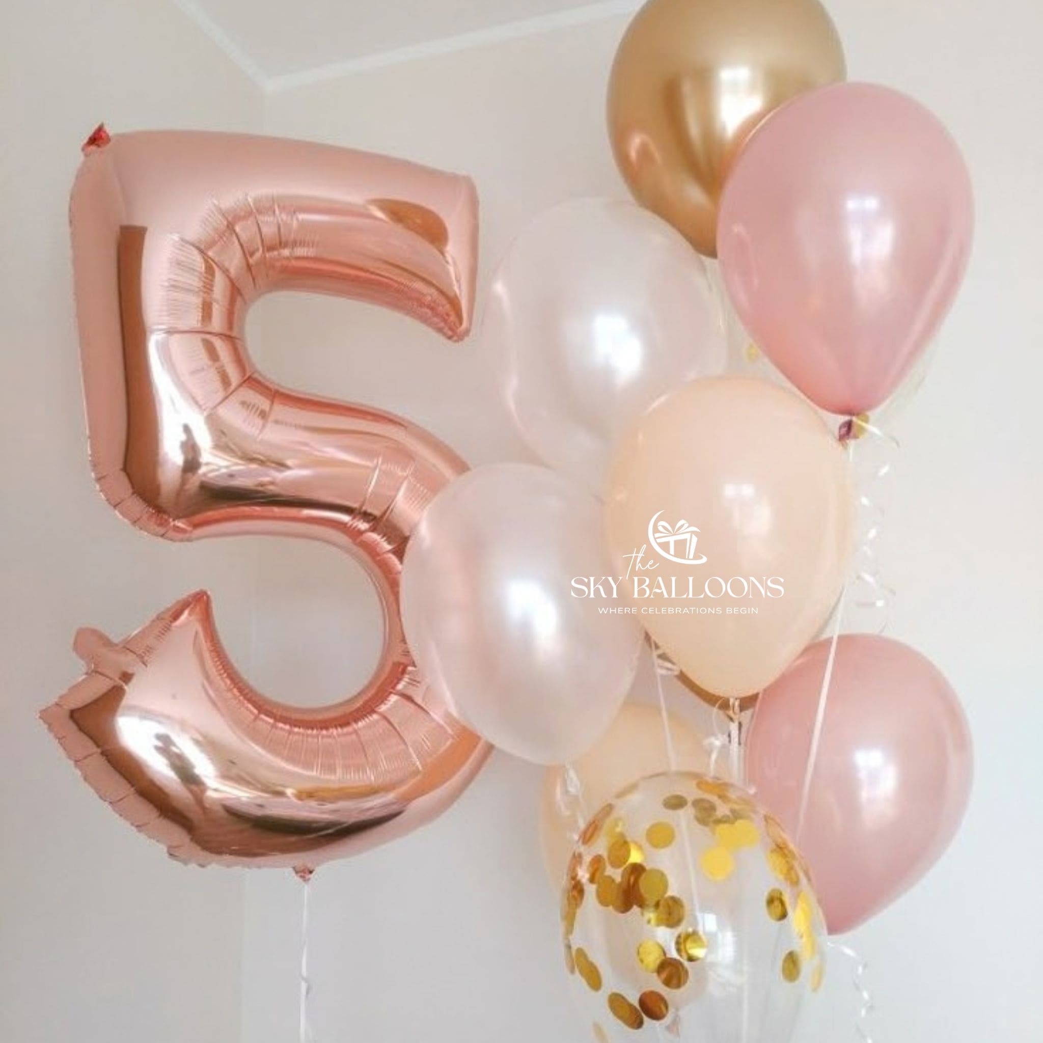 A cluster of metallic and latex balloons, including a large number 5 foil balloon in rose gold, several blush pink balloons, one champagne gold balloon, and a clear balloon filled with gold confetti, against a plain white wall.