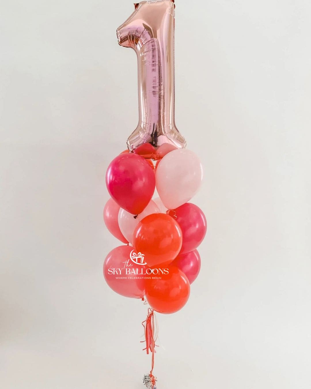 A first birthday balloon bouquet featuring a metallic rose gold foil balloon shaped like the number '1' at the top, with a cluster of round balloons in shades of pink, red, and light pink below. The balloons are tied together and hang against a plain white background.