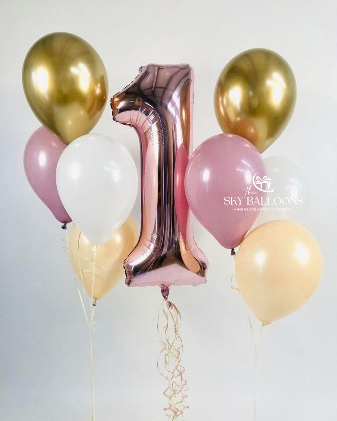 A cluster of metallic and pastel balloons, including a large foil number 1 balloon in rose gold, gold, pink, white, pale yellow, and blush pink latex balloons, tied together with a string against a light gray background. 