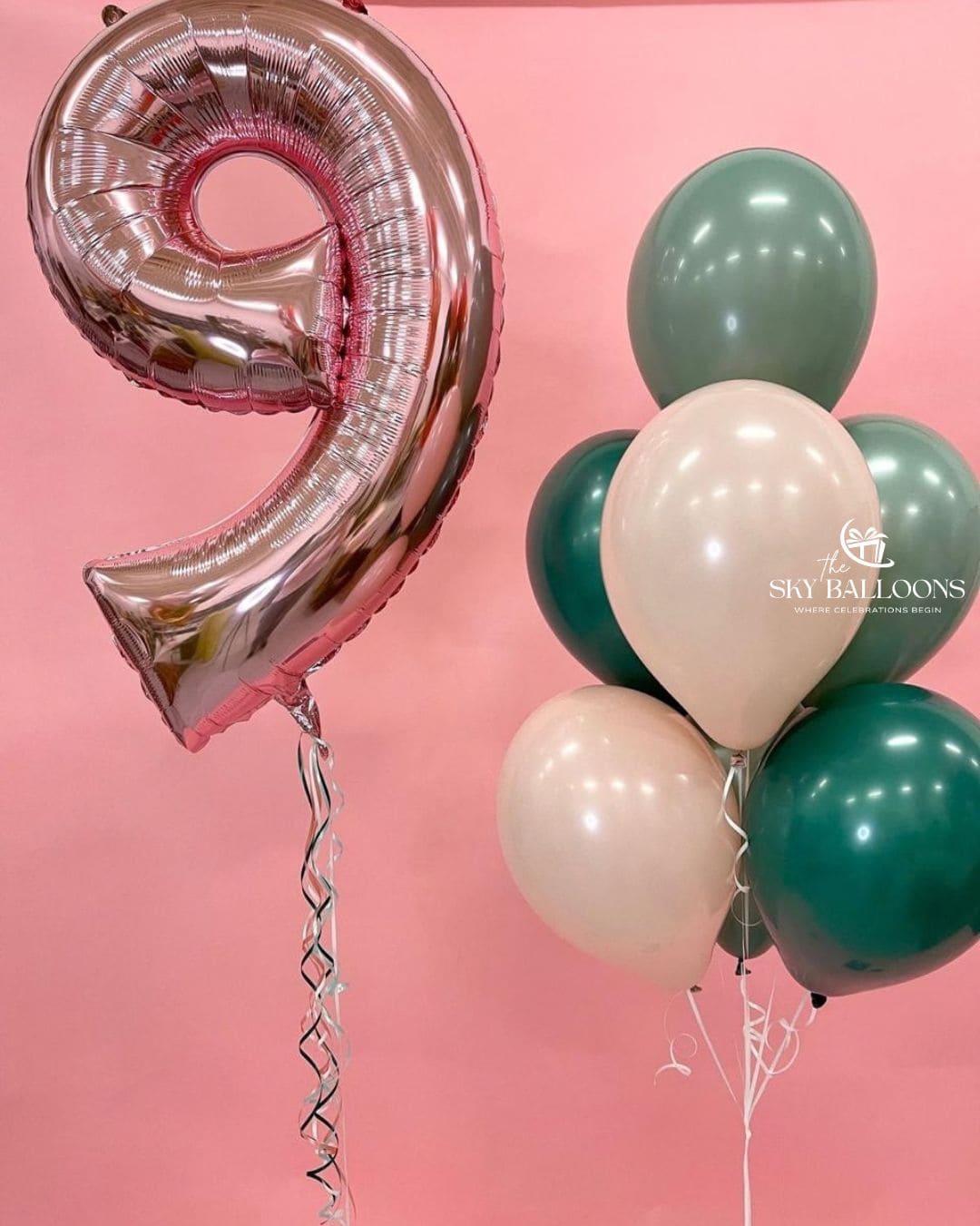 Rose gold number 9 foil balloon beside a bouquet of sage green, dark green and cream latex balloons against a pink background.