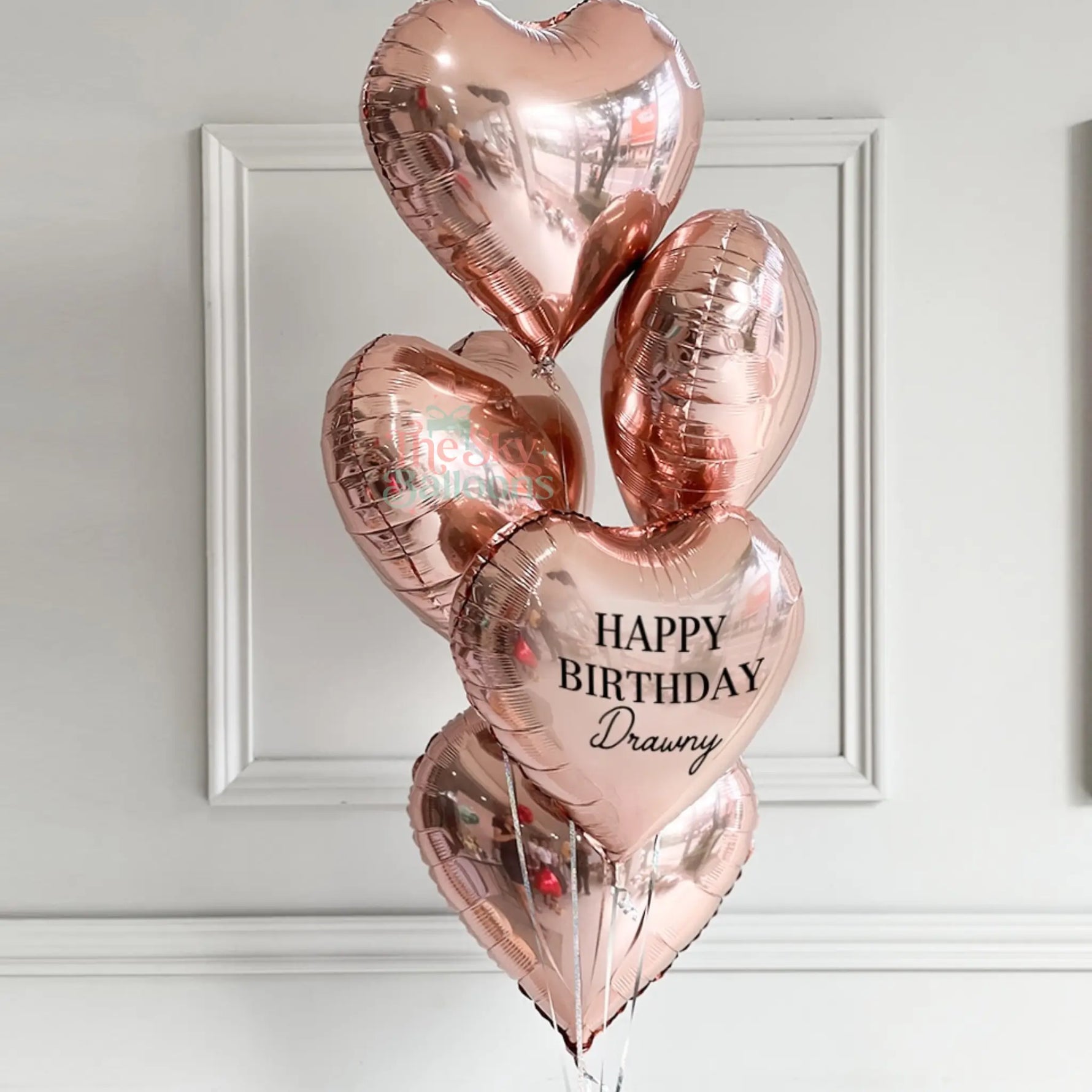 Rose gold heart-shaped balloons with 'Happy Birthday Drawny' text on a white wall background