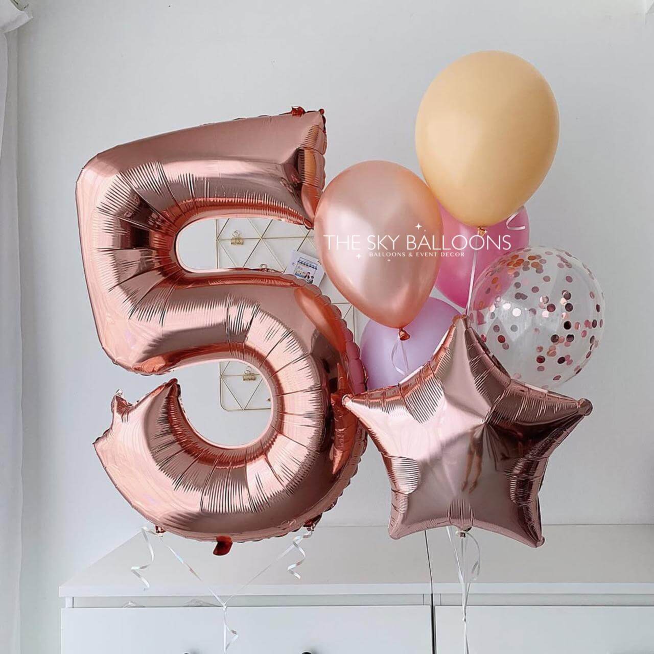 Rose gold number '5' balloon with other balloons against a white wall.