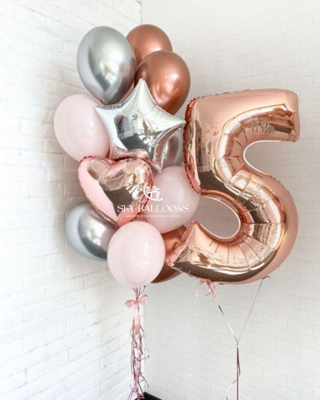 A cluster of metallic and pastel balloons, including a large rose gold number 5 foil balloon, a silver star balloon, a pink heart balloon, and several round balloons in rose gold, silver, and light pink, tied together and photographed against a white brick wall.