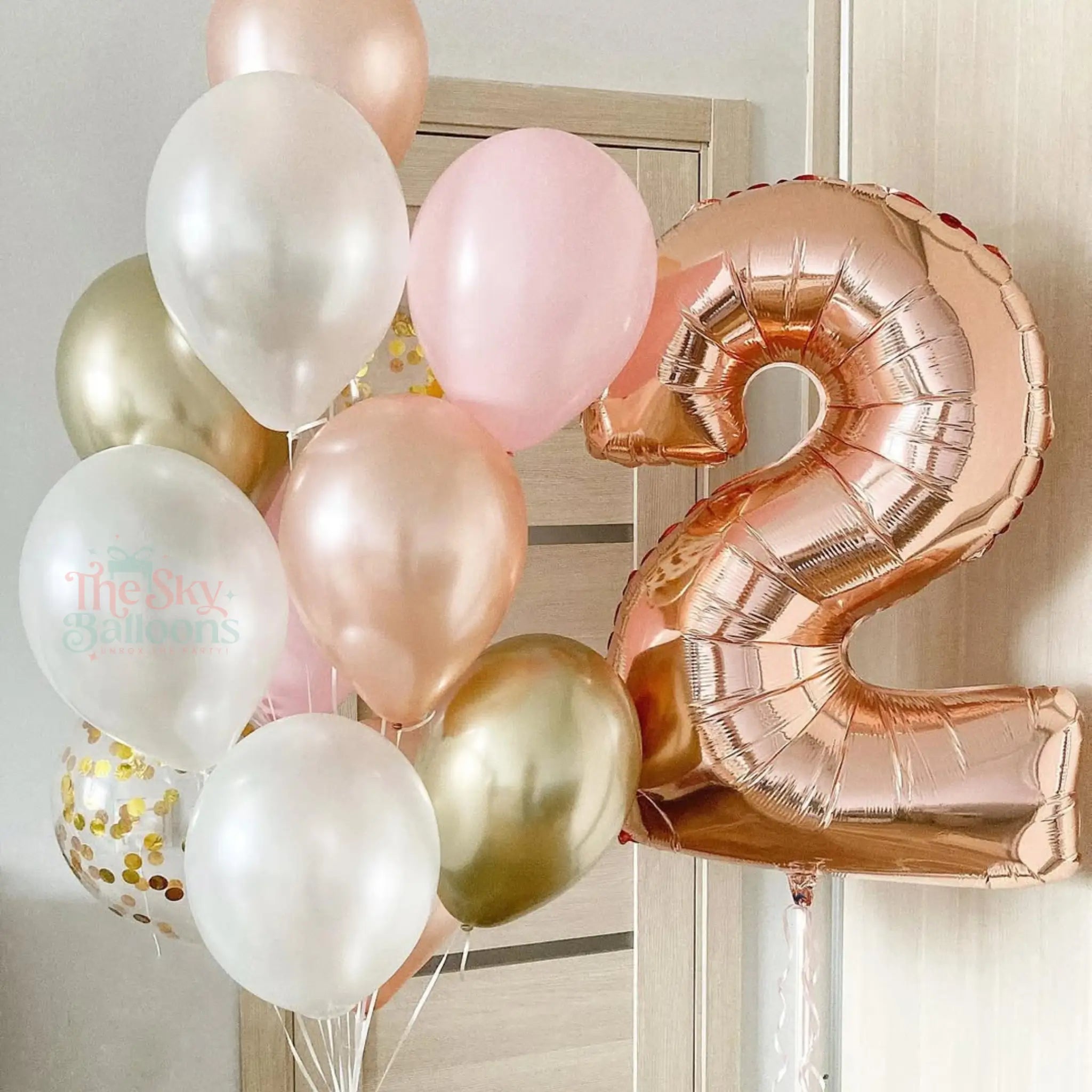 Rose gold, pink, and white balloons with a number 2 balloon against a neutral wall.