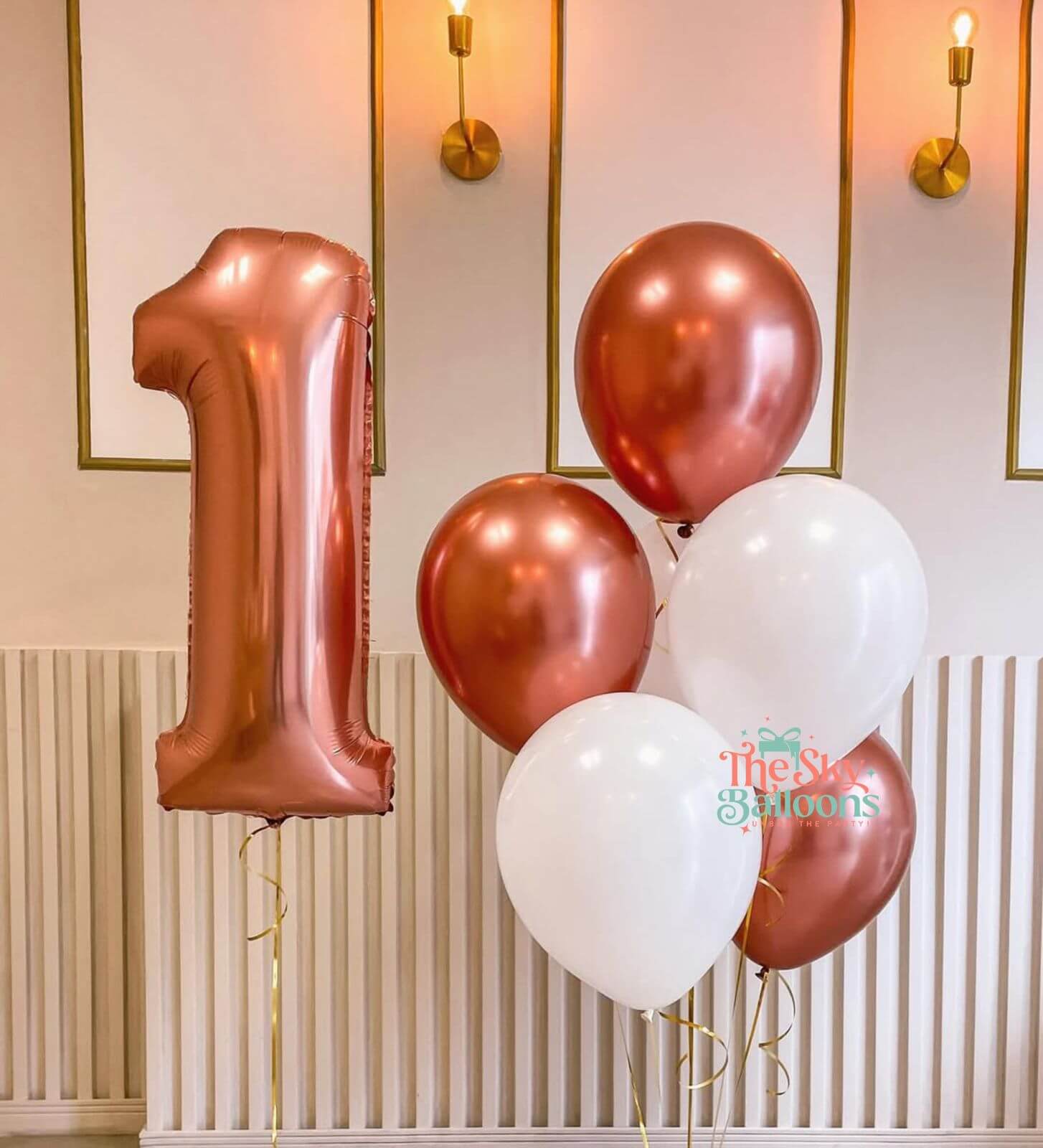 Rose gold and white balloons with a number 1 balloon against a decorative wall.