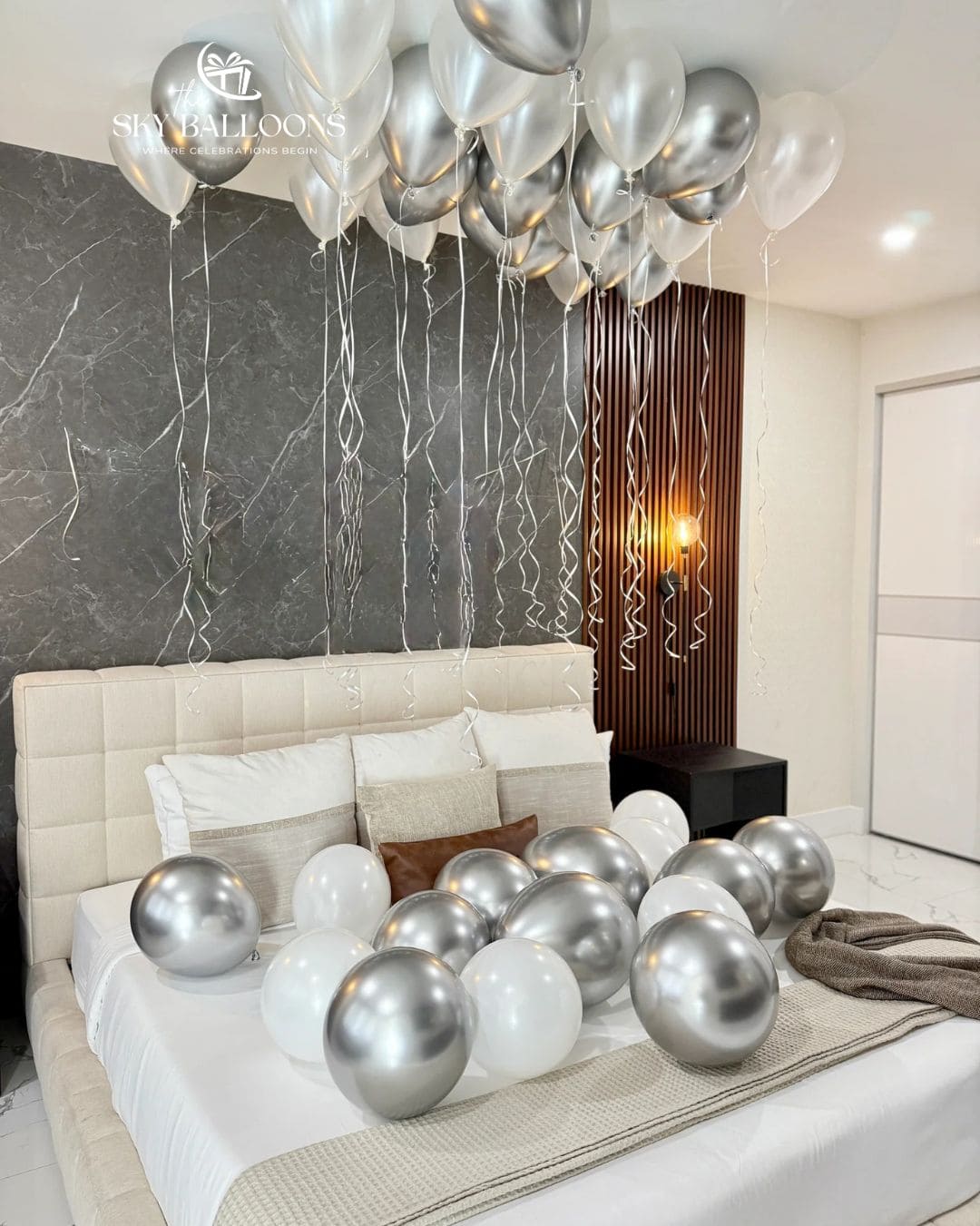 Bedroom decorated with metallic silver and pearl white ceiling balloons with long curling ribbons and matching balloons scattered across the bed.