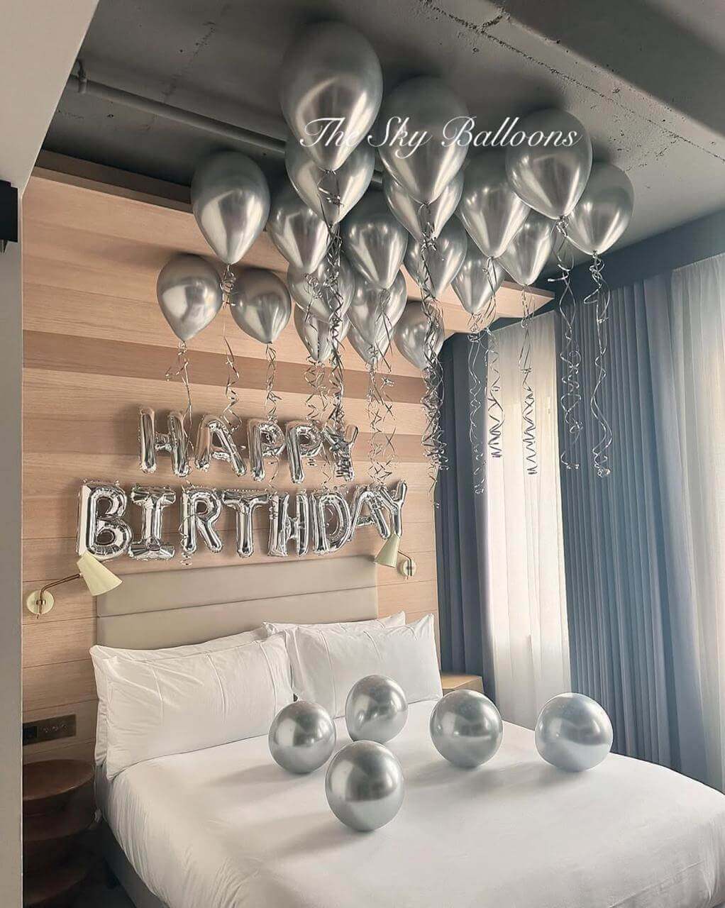 Sophisticated silver birthday balloon decoration featuring 'HAPPY BIRTHDAY' letter balloons with matching silver helium balloons floating on ceiling and chrome balloons arranged on white hotel bed in modern bedroom with wood accent wal