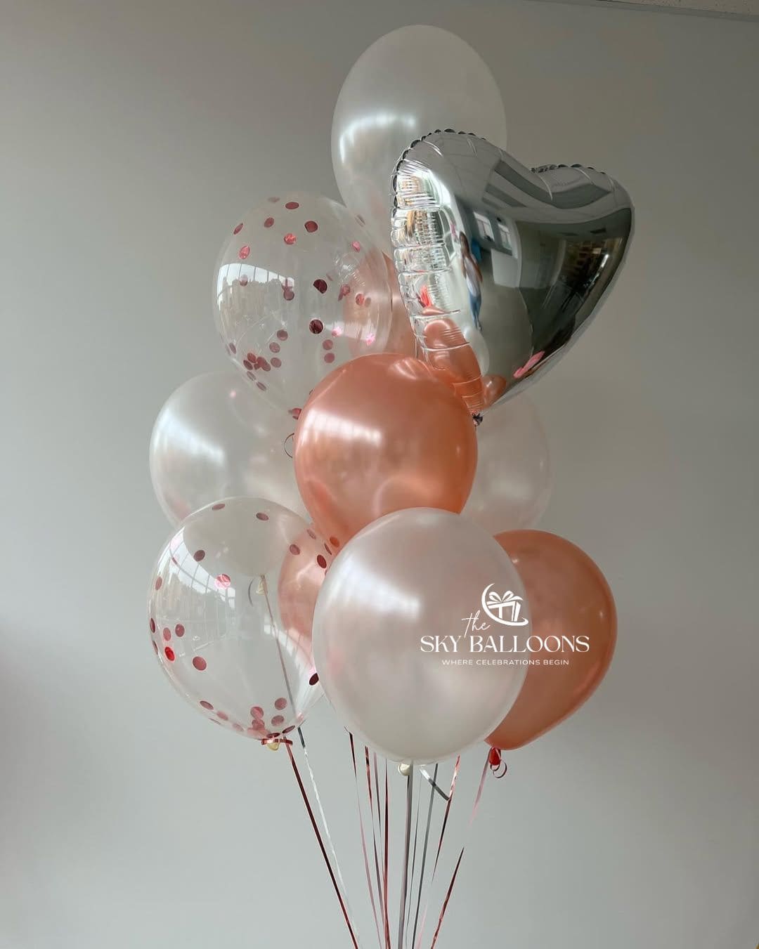A cluster of party balloons including clear balloons with red confetti, solid peach and white balloons, and a metallic silver heart-shaped balloon, tied together with red strings against a plain light gray wall.