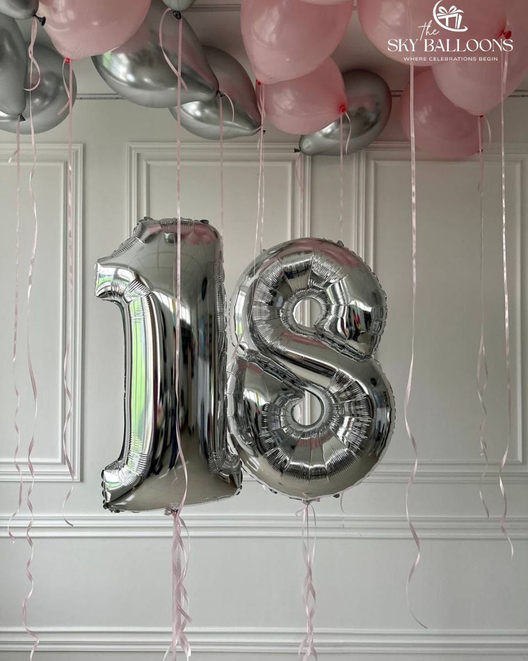 Silver number 18 foil balloons beneath blush pink and metallic silver ceiling balloons with long pale pink ribbons against a white panelled wall.