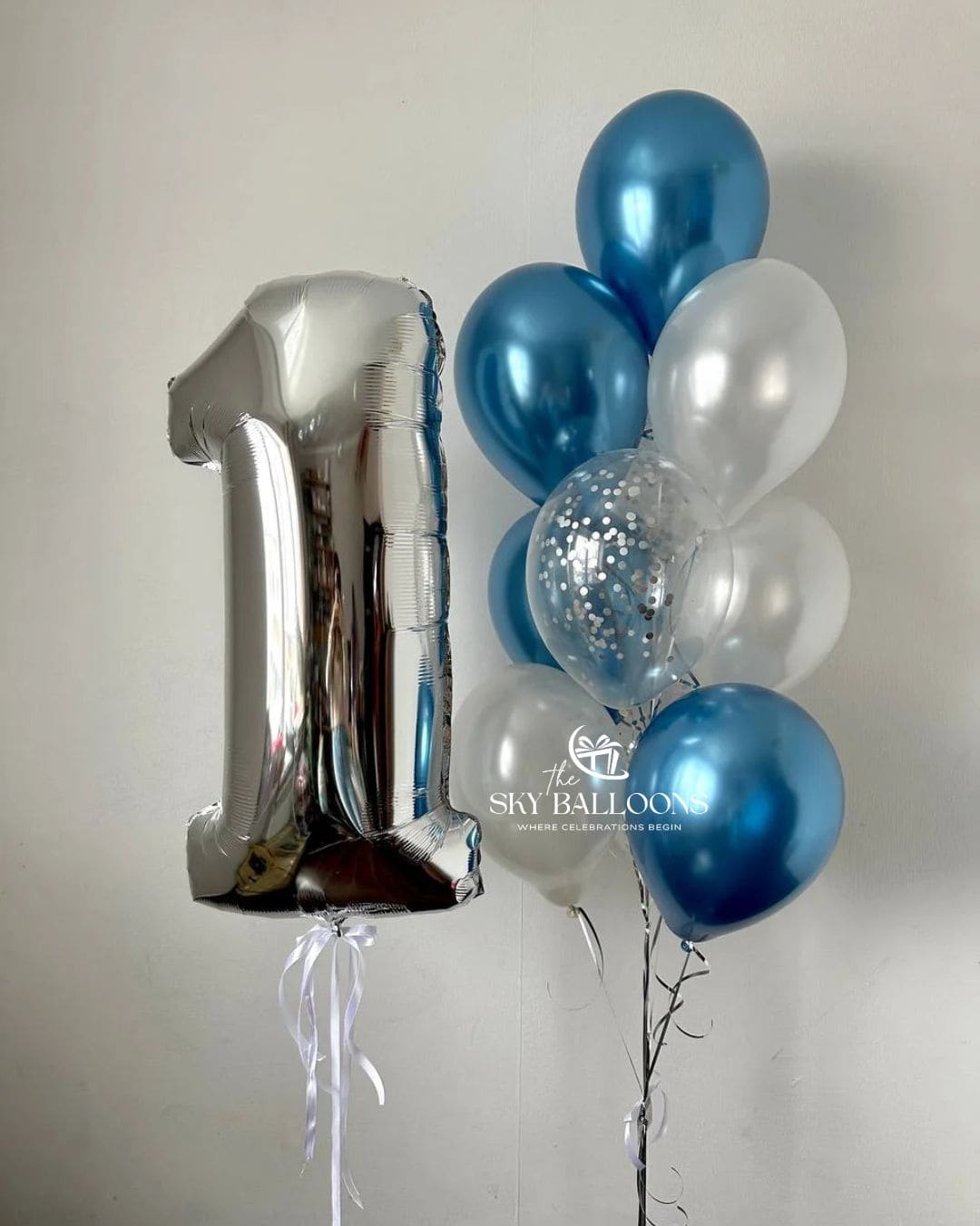 A silver foil balloon shaped like the number 1 stands next to a cluster of metallic blue, white, and clear confetti balloons tied to a black stand against a plain light gray wall. 