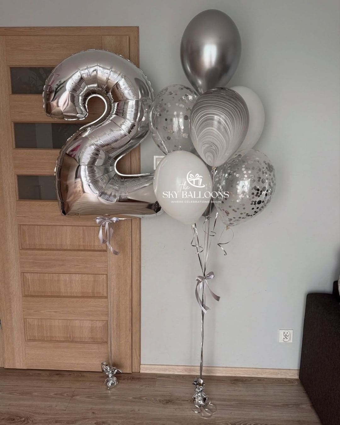 A cluster of metallic silver and white balloons, including a large number 2 foil balloon and several round balloons, tied to a floor stand in an indoor room with a wooden door and light gray wall.