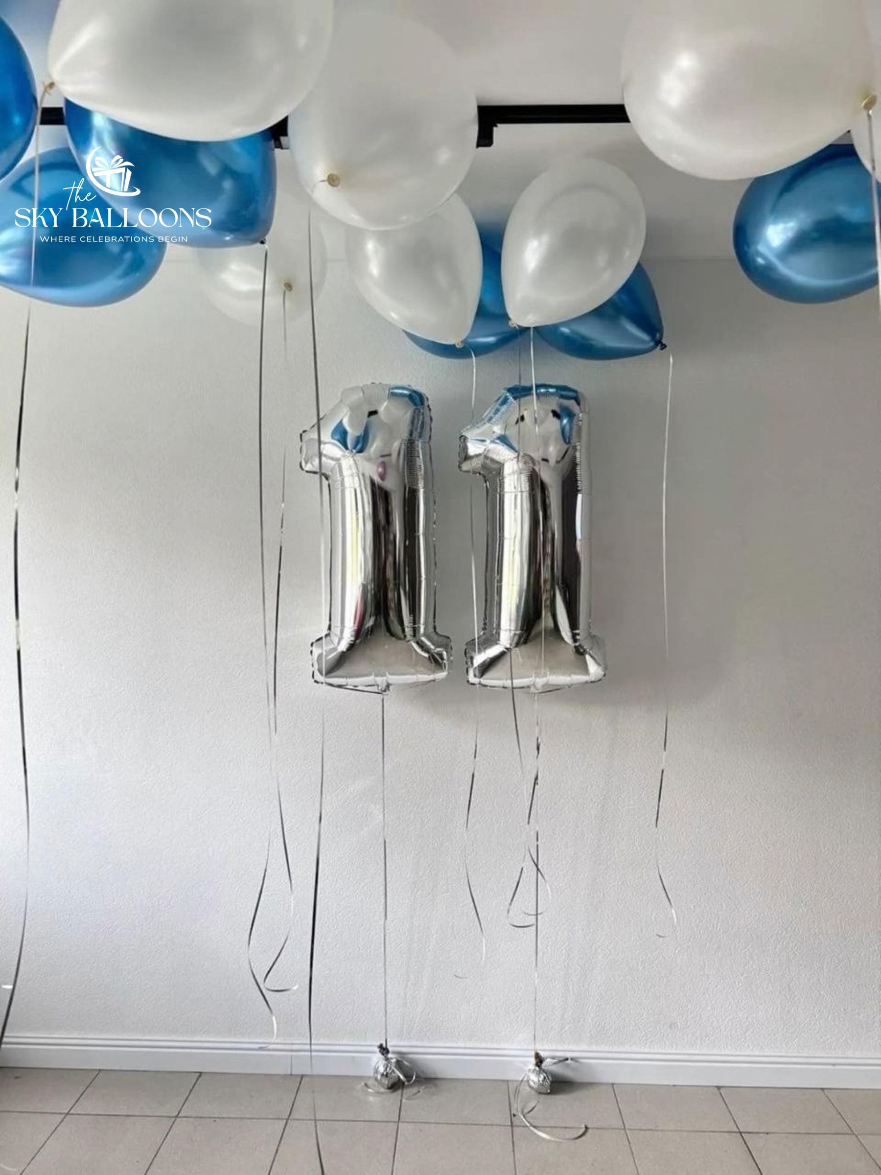 Two metallic silver foil balloons shaped like the number 11, hanging from a black ceiling bar with white and blue balloons above them, against a white wall.