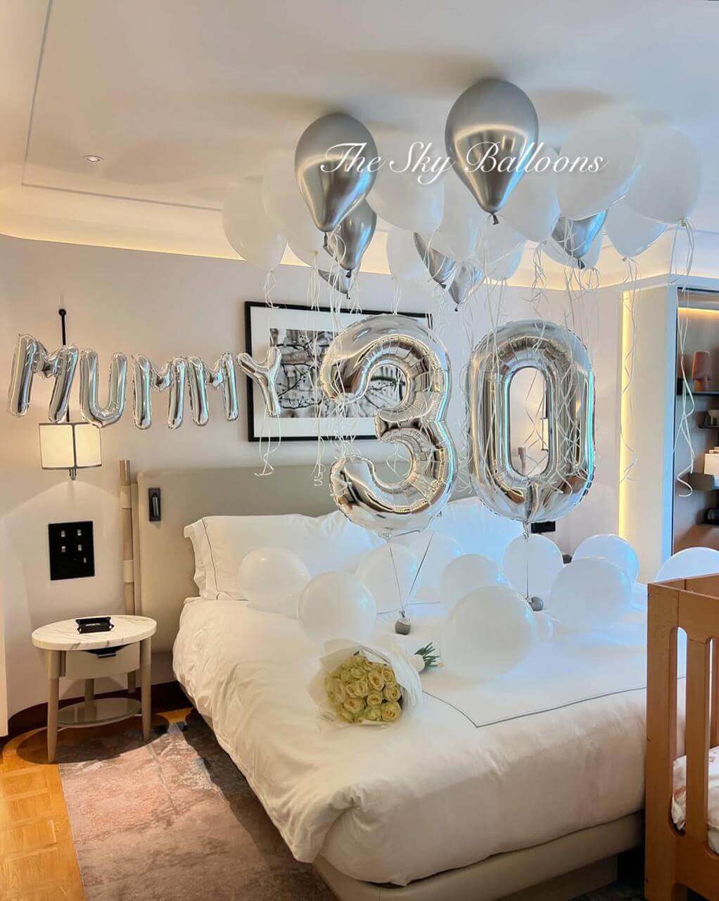 Elegant 30th birthday balloon decoration featuring silver 'MUM' letter balloons and large silver foil number '30' balloons with white and silver helium balloons floating on ceiling, displayed in modern hotel bedroom with white roses