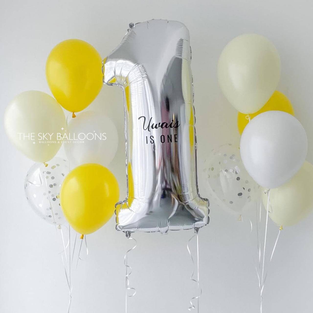 Silver balloon shaped like the number '1' with 'Winnie is one' text, surrounded by yellow and white balloons on a light gray background.