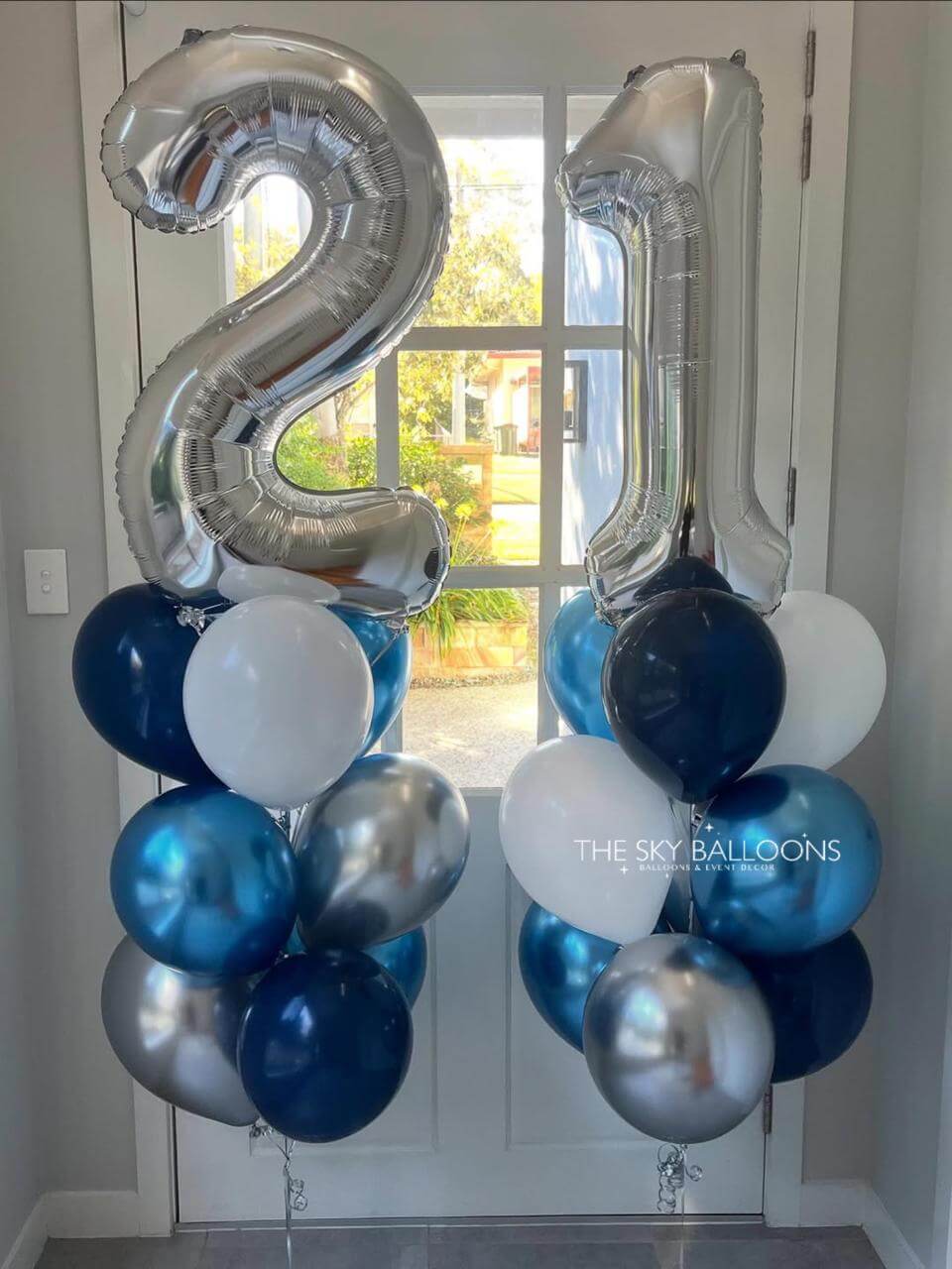 Silver, blue, and white balloons shaped like the number '21' in a room with a window.
