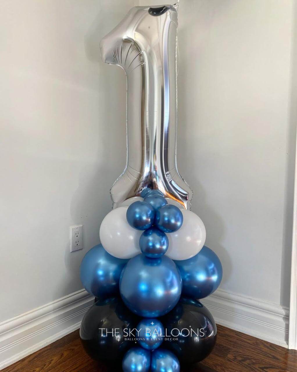 Silver number '1' balloon with blue and white balloons against a plain wall.