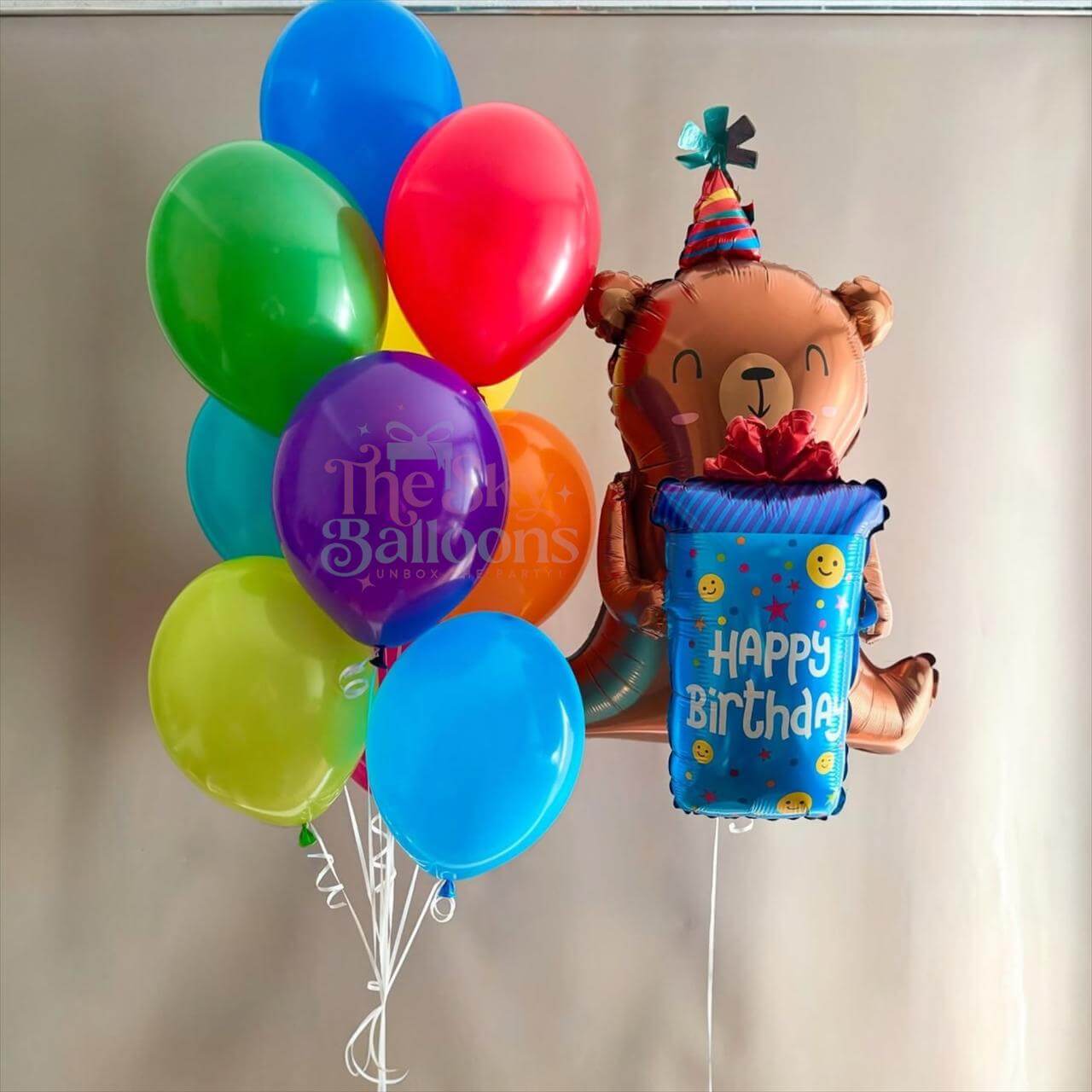Colorful balloons with a bear-shaped balloon holding a 'Happy Birthday' box against a plain background.
