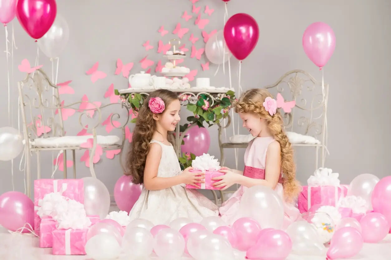 Two young girls in a decorated room with pink balloons and gifts, holding a gift box.
