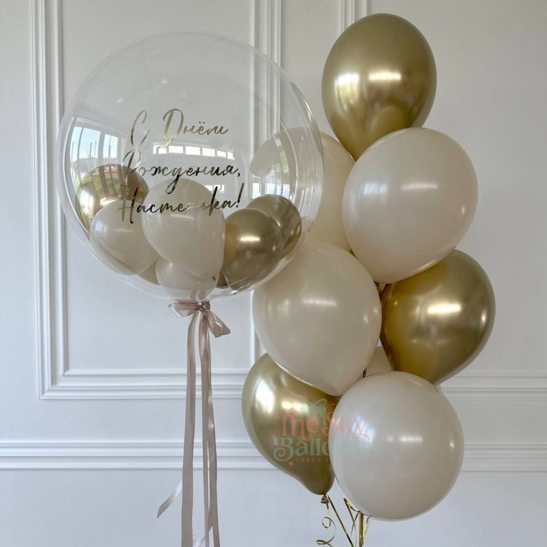 Bouquet of gold, white sand, and clear balloons with visible branding against a white wall.