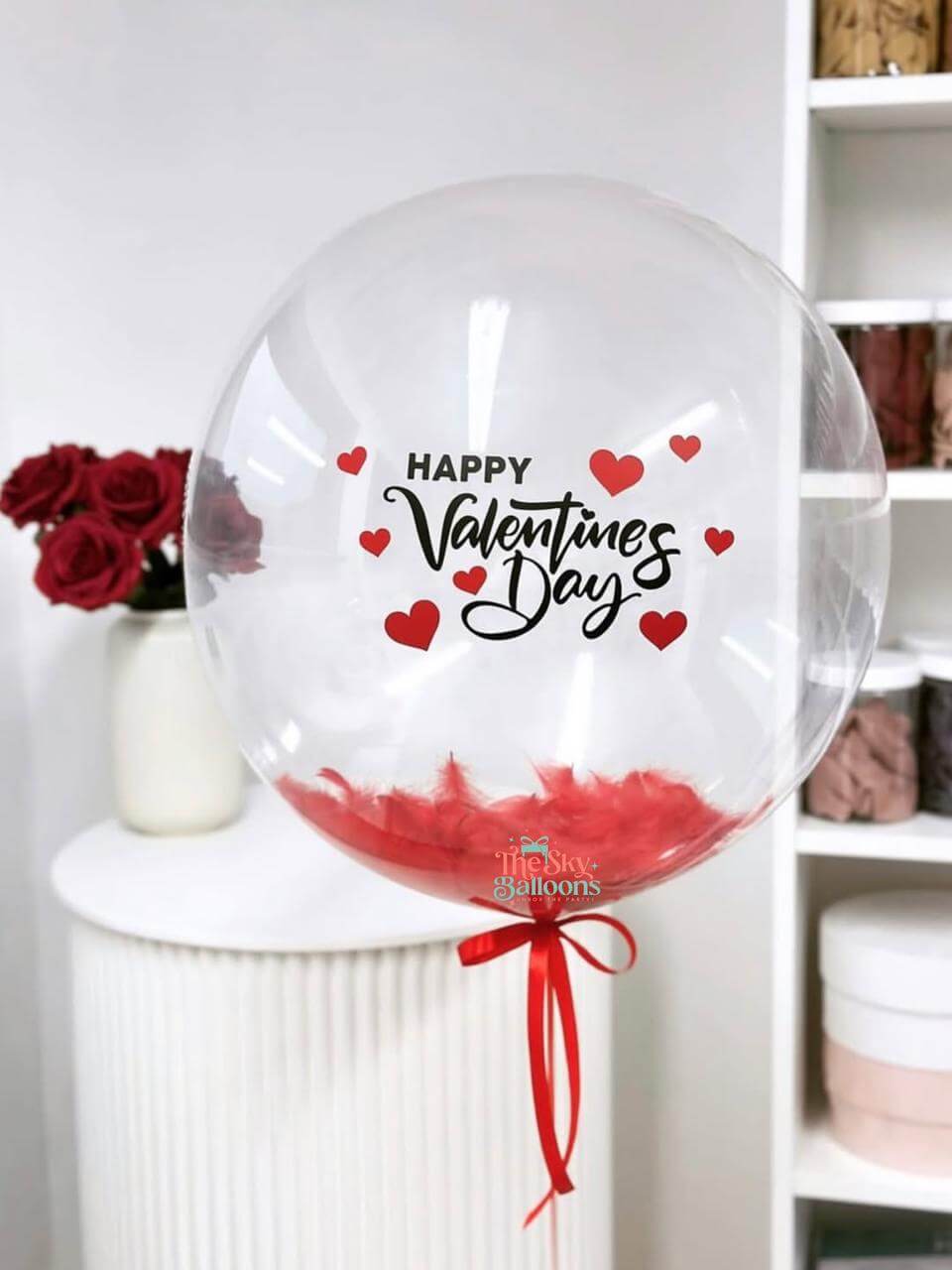 Clear balloon with 'Happy Valentine's Day' text and red hearts, tied with a red ribbon, on a white surface.