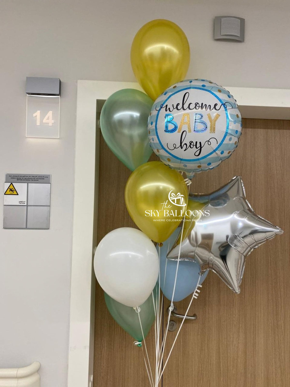 A cluster of baby shower balloons including a round foil balloon that reads 'welcome BABY boy' with blue polka dots, metallic gold, white, light blue, and silver balloons, tied next to a wooden door.