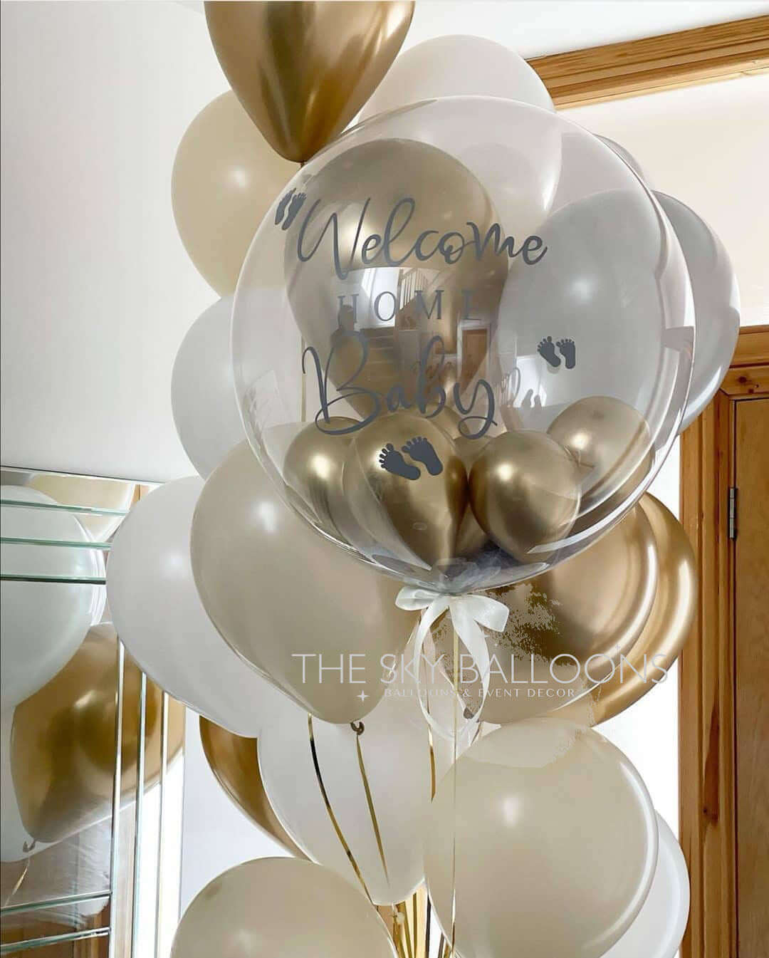 A bouquet of latex balloons in shades of gold, white, and transparent, with 'Welcome Home' text printed on a large gold balloon, tied with ribbons and helium-filled.