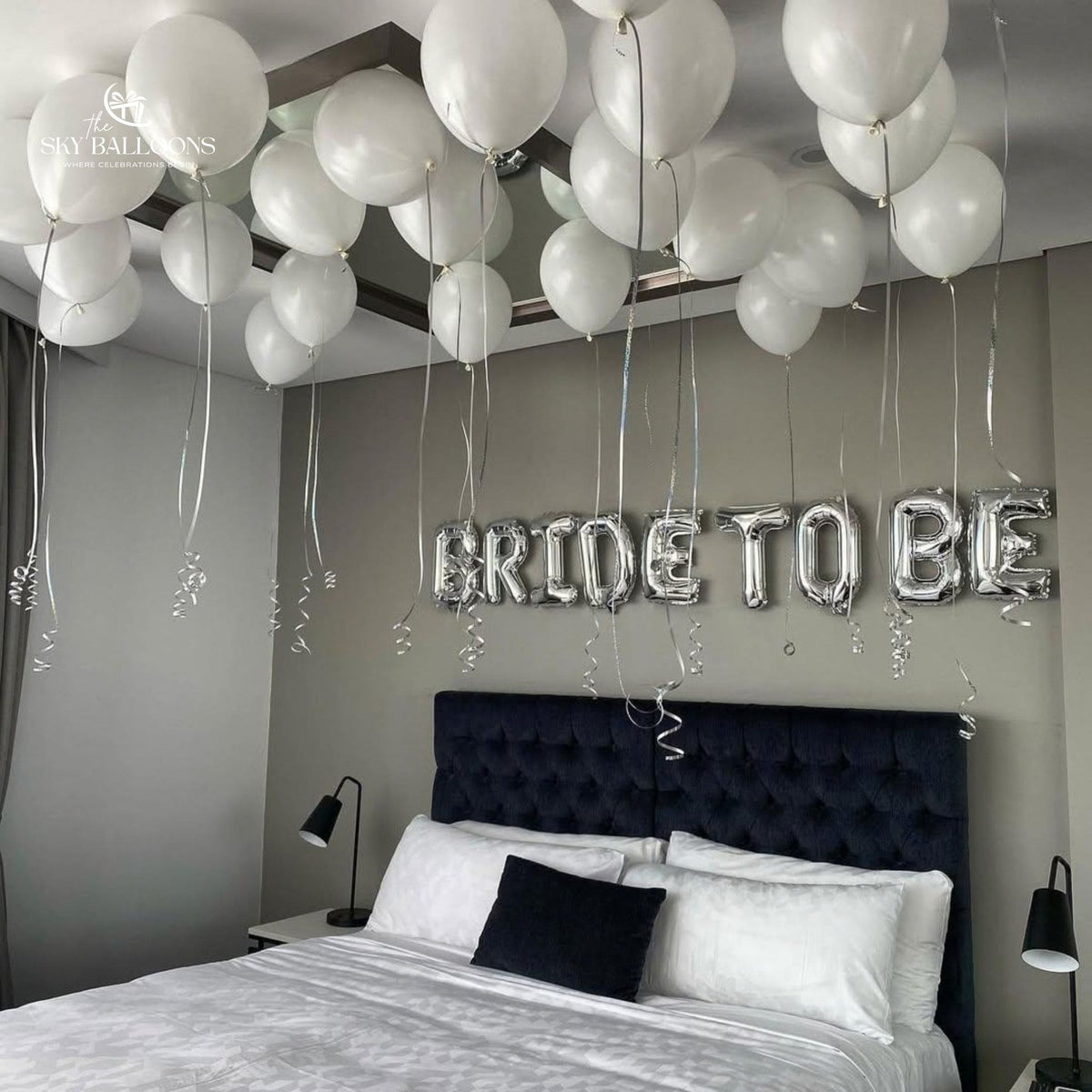 White ceiling balloons with hanging silver ribbons and a silver Bride To Be foil banner above a bed.