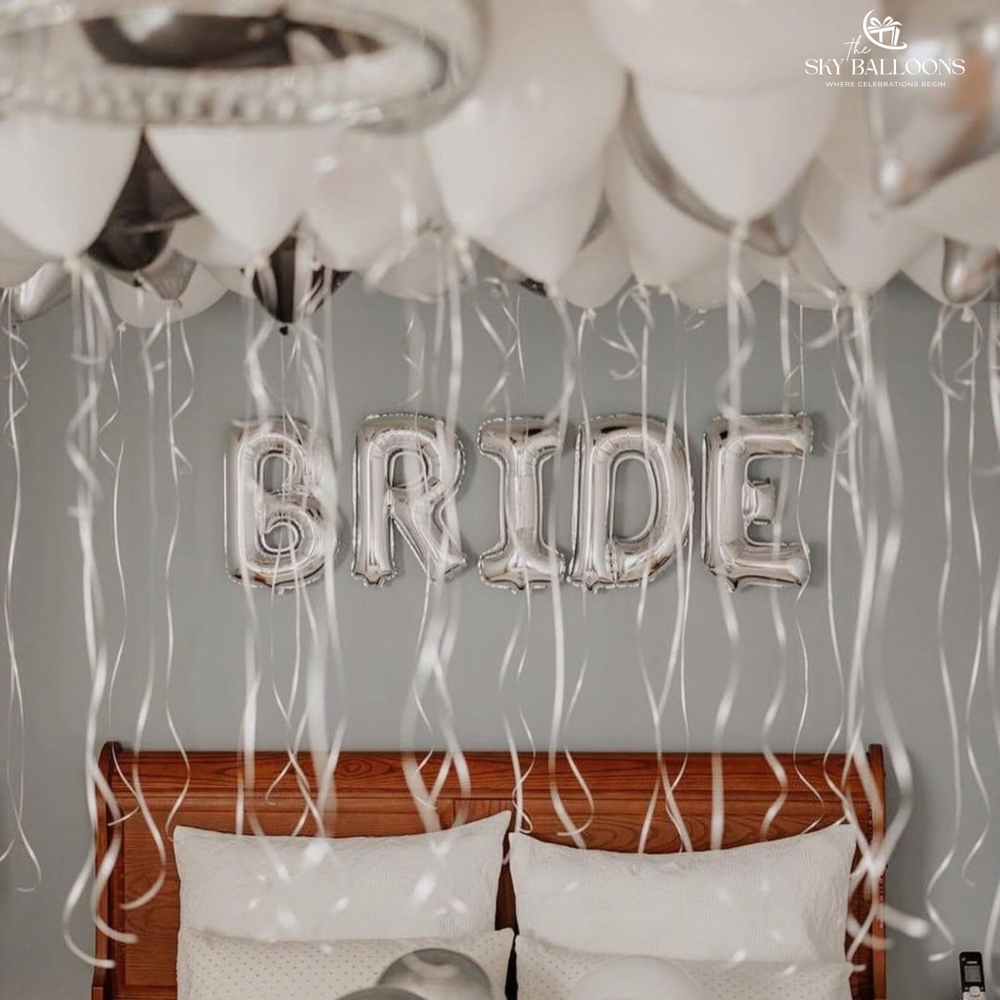 White and silver ceiling balloons with hanging ribbons and a silver BRIDE foil banner above a bed.