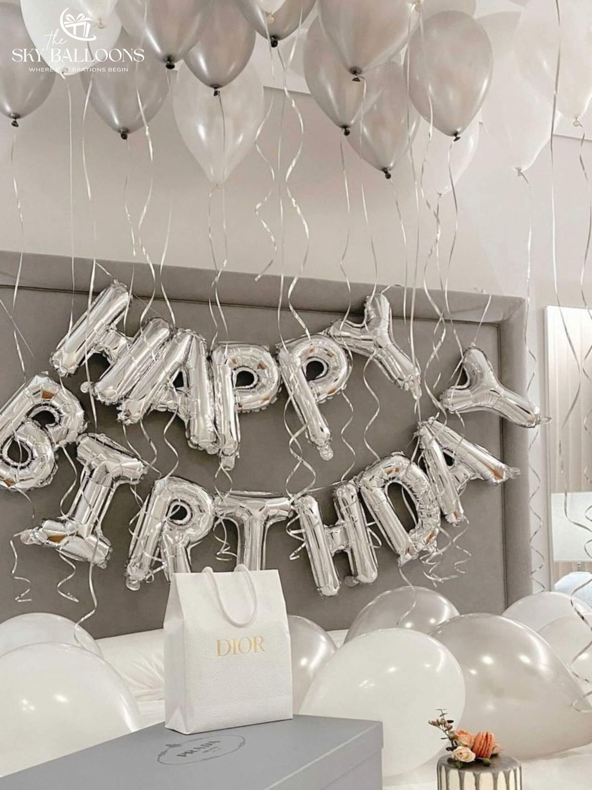 White and pearl-silver ceiling and floor balloons with curling ribbons and a silver Happy Birthday foil banner above a bed.