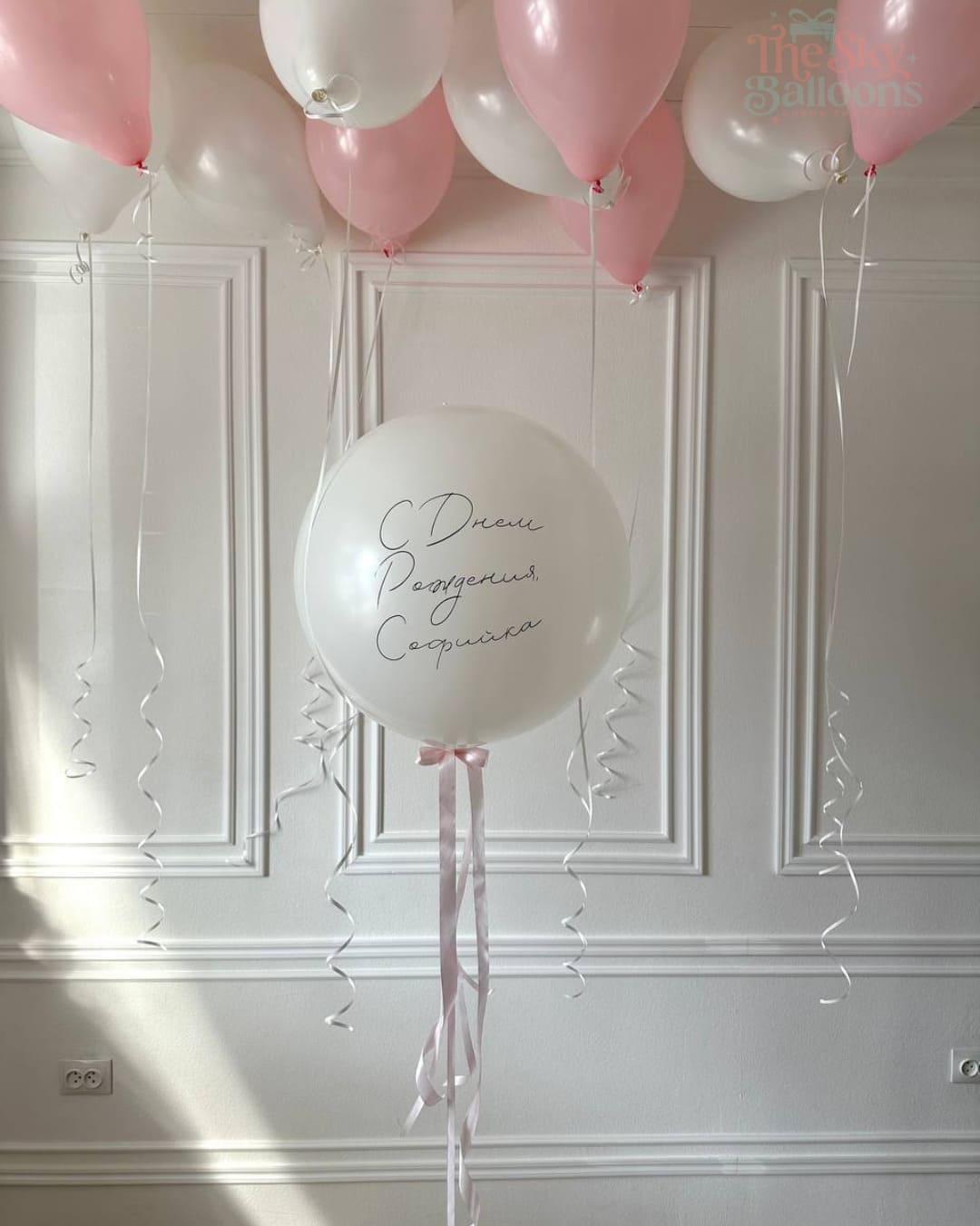 White balloon with cursive text against a white paneled wall with pink and white balloons.