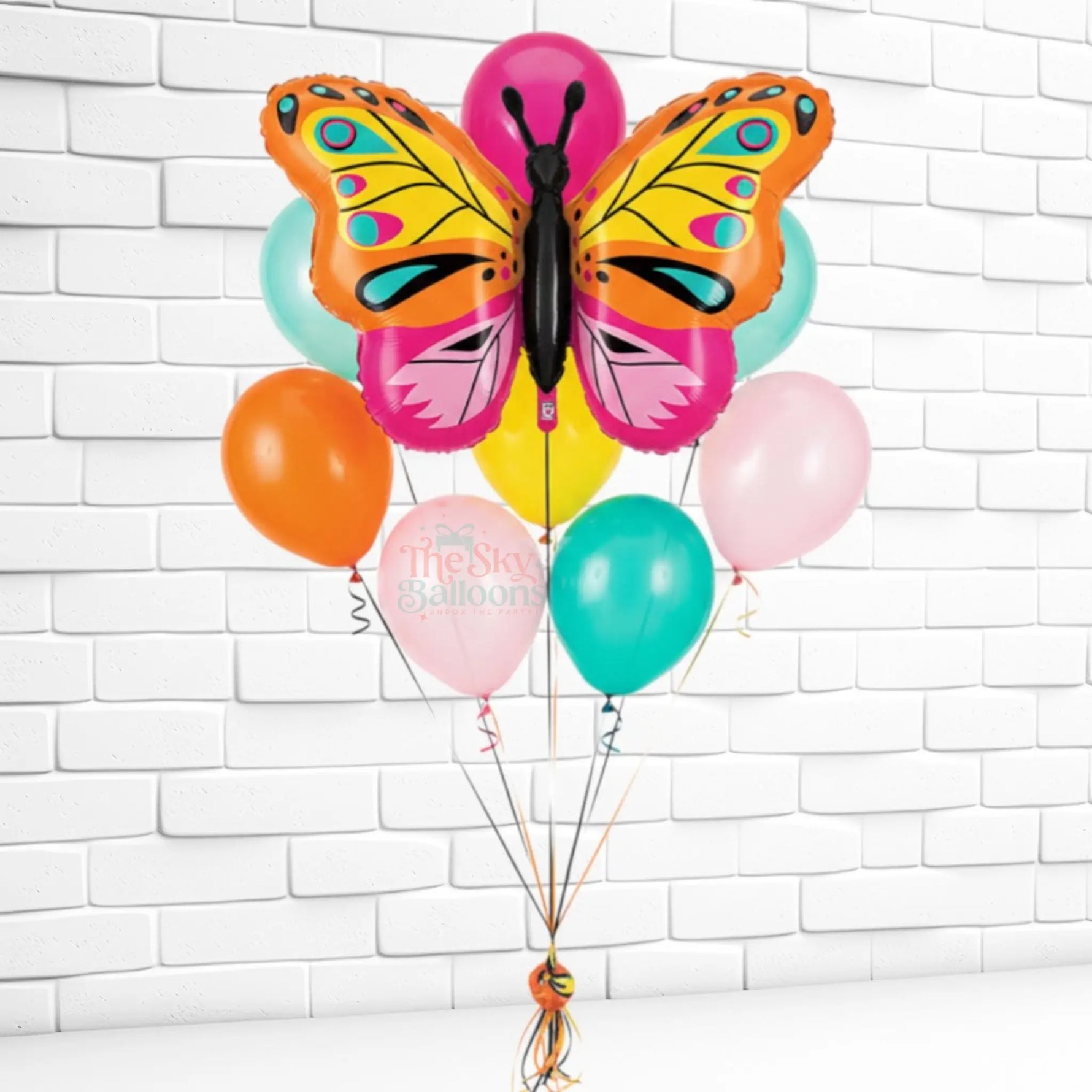 Colorful butterfly-shaped balloon with other balloons against a white brick wall.