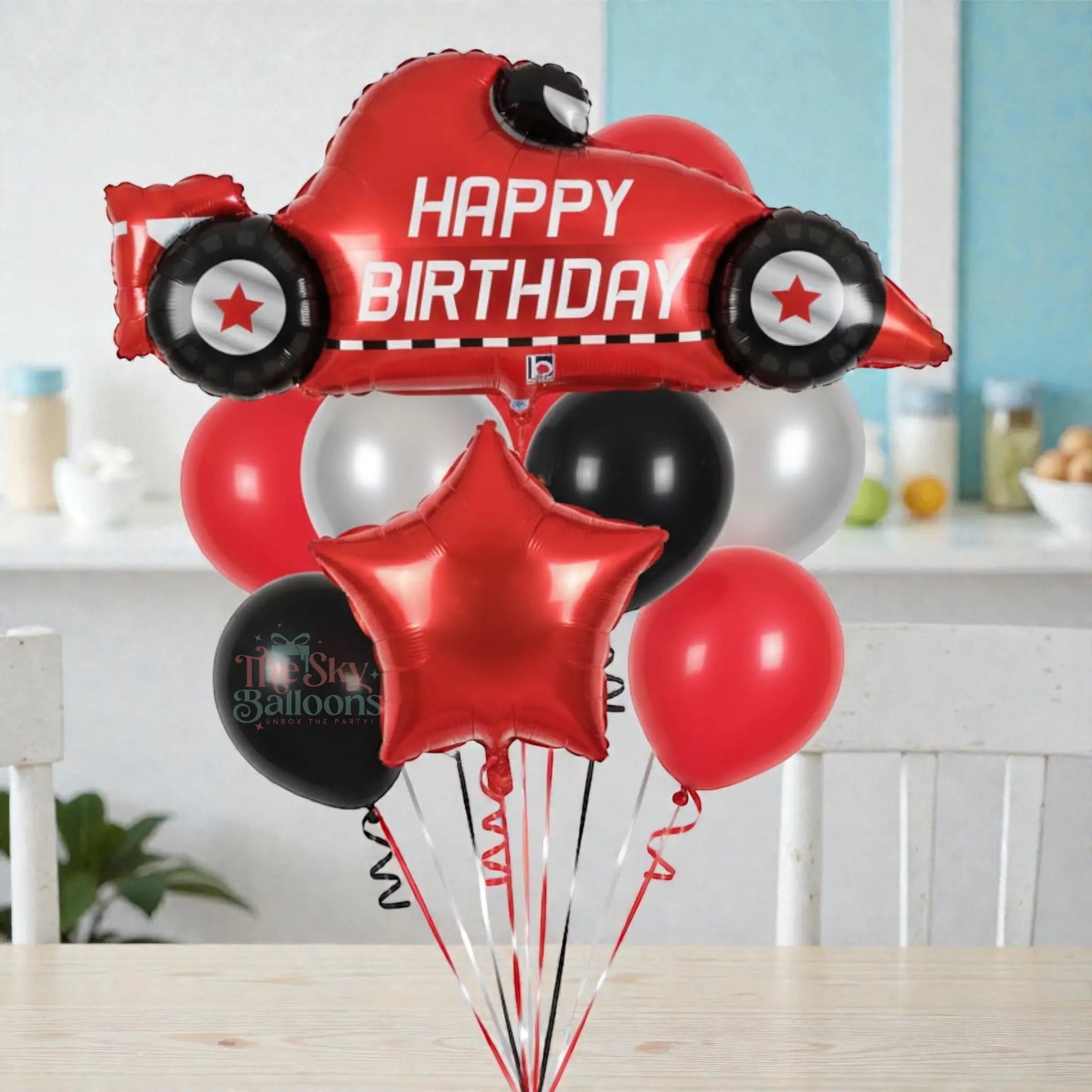 Red race car-shaped balloon with 'Happy Birthday' text, surrounded by other balloons, held against a white background.