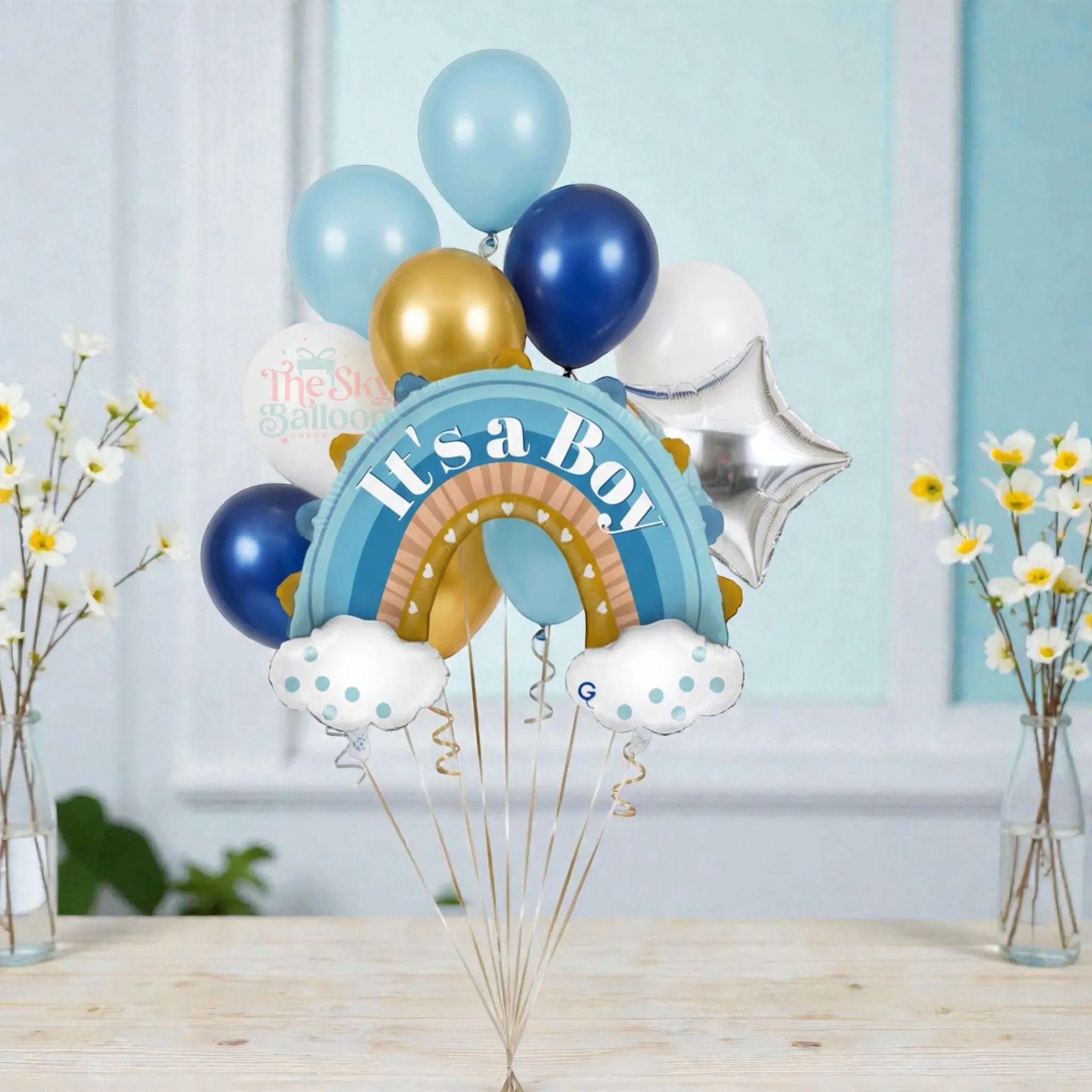 Balloon bouquet with 'It's a Boy' message on a stand, featuring blue, gold, and silver balloons against a light blue wall.