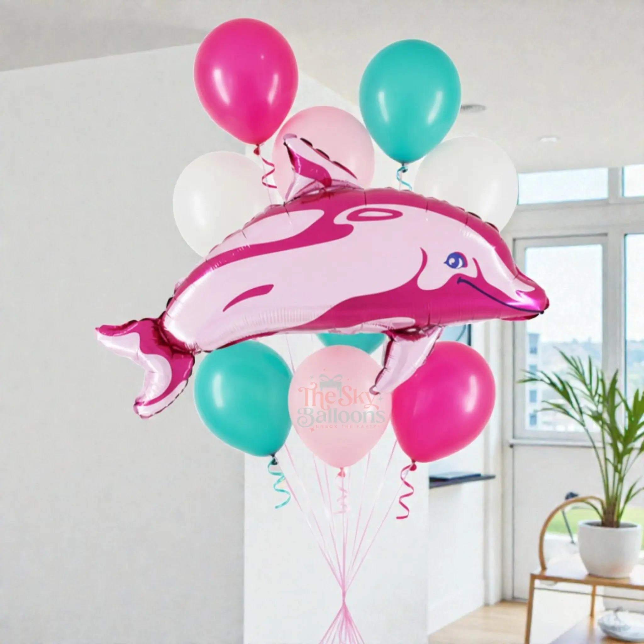 Pink dolphin-shaped balloon among other balloons in a room with a plant and window.