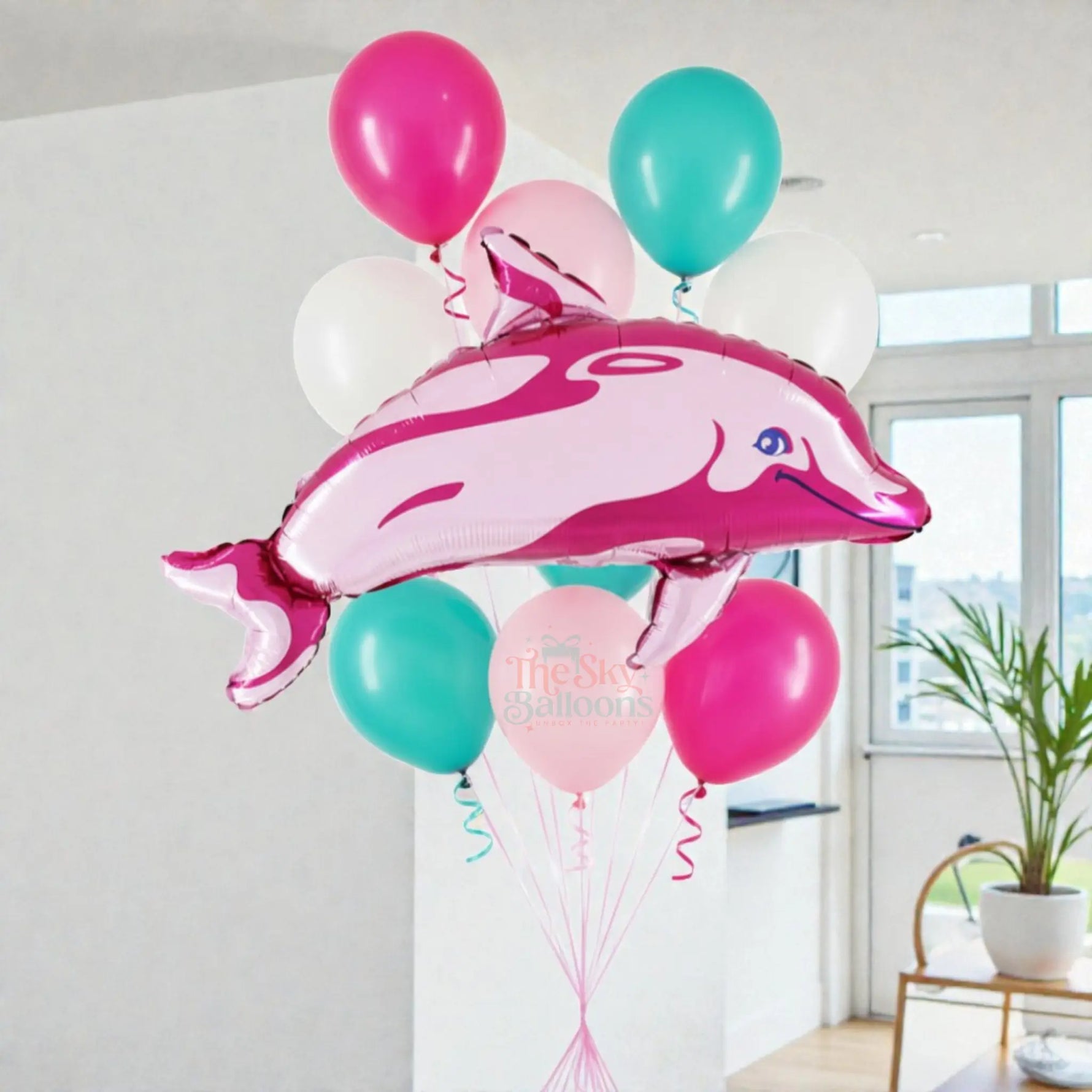 Pink dolphin-shaped balloon among other balloons in a room with a plant and window.