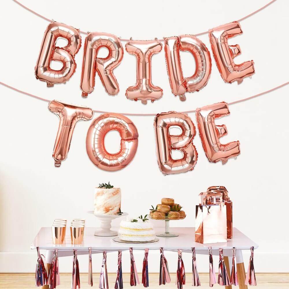 Rose gold 'BRIDE TO BE' balloons above a table with a cake and drinks.