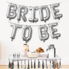Silver 'BRIDE TO BE' balloons above a table with a cake and snacks.