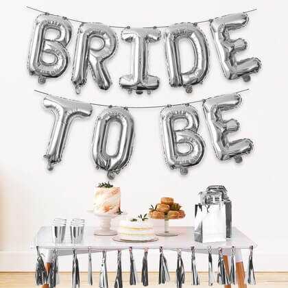 Silver 'BRIDE TO BE' balloons above a table with a cake and snacks.