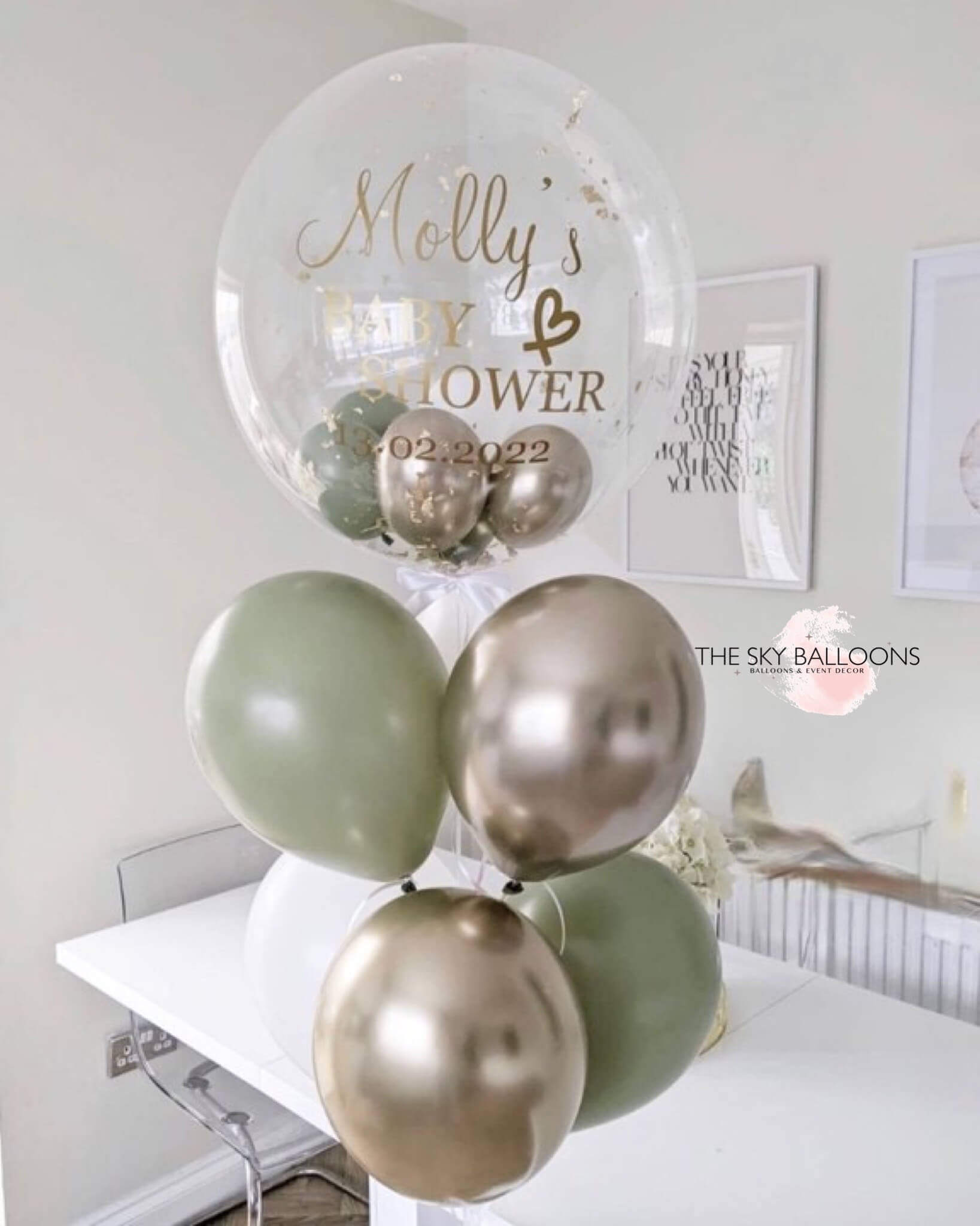 A bouquet of balloons including eucalyptus, white, and chrome gold colors, with a personalized jumbo balloon that reads 'Molly's Baby & Hair' with a date.