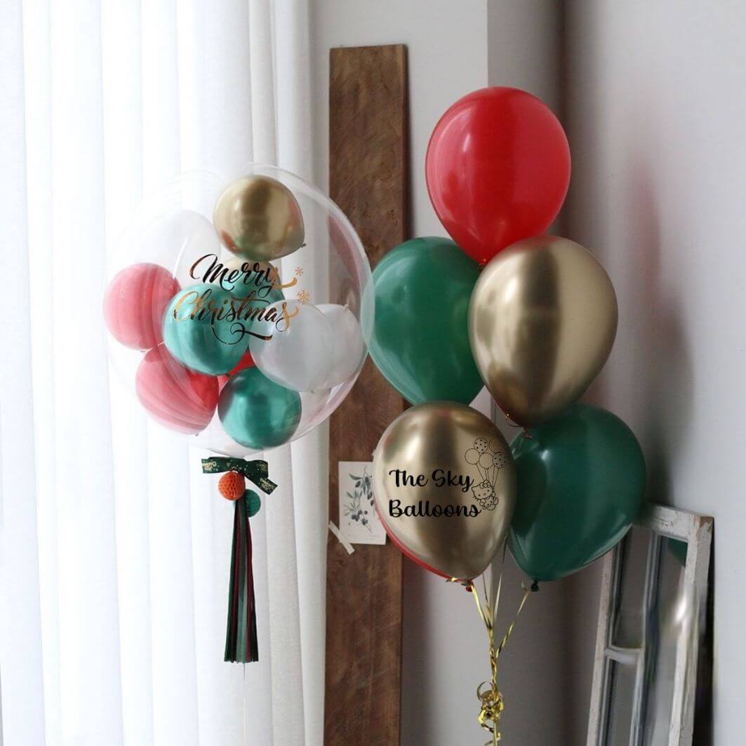 A bouquet of round latex balloons in various colors including gold, green, red, and white, with 'Merry Christmas' and other festive messages written on them, tied with ribbons and hanging against a wall.
