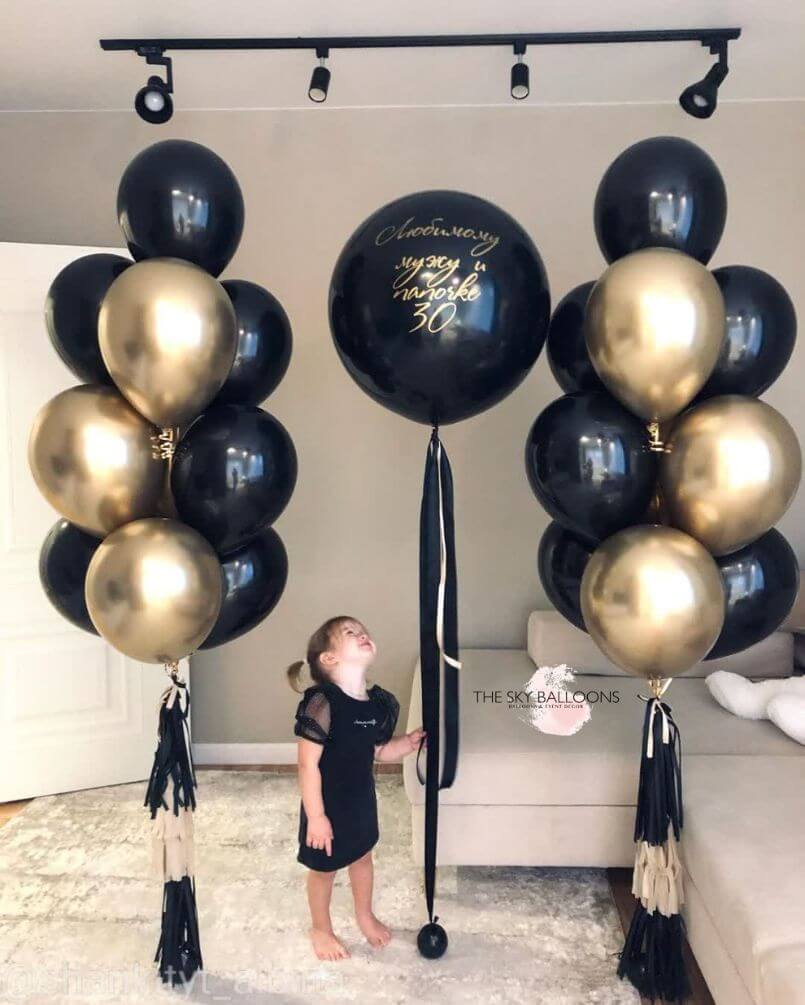 A child standing beside a bouquet of black and gold balloons with a customized balloon that has 'Anniversary' written on it.