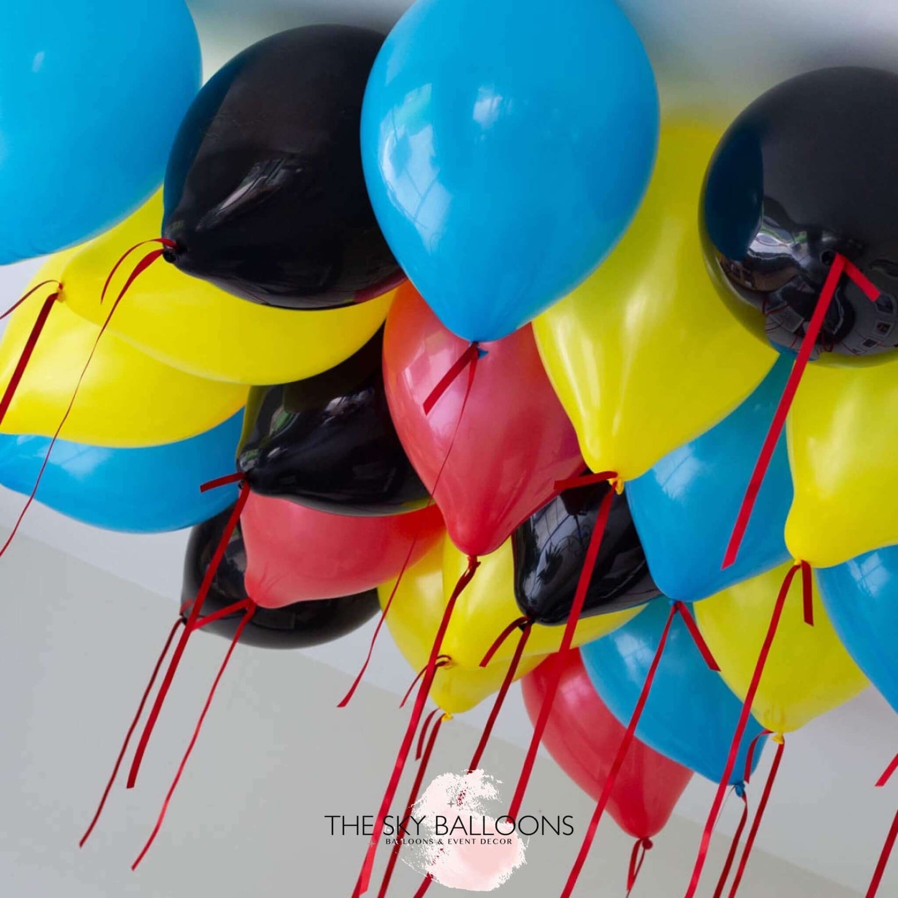 A cluster of inflated ceiling balloons in red, blue, yellow, and black colors with ribbons attached, delivered by The Sky Balloons.