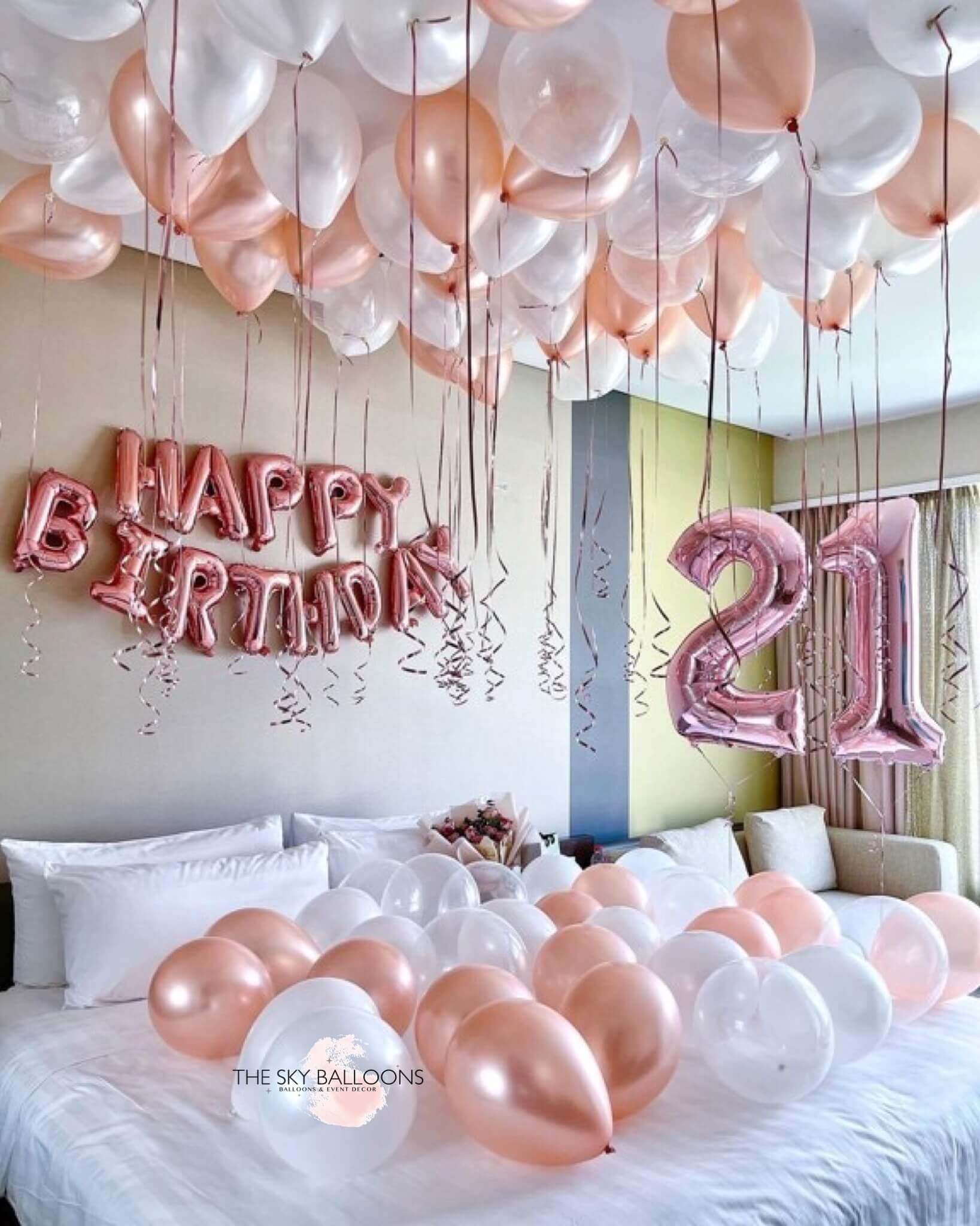 A room decorated with rose gold and pearl white balloons, including a 'Happy Birthday' banner and large numbered balloons spelling '21'.
