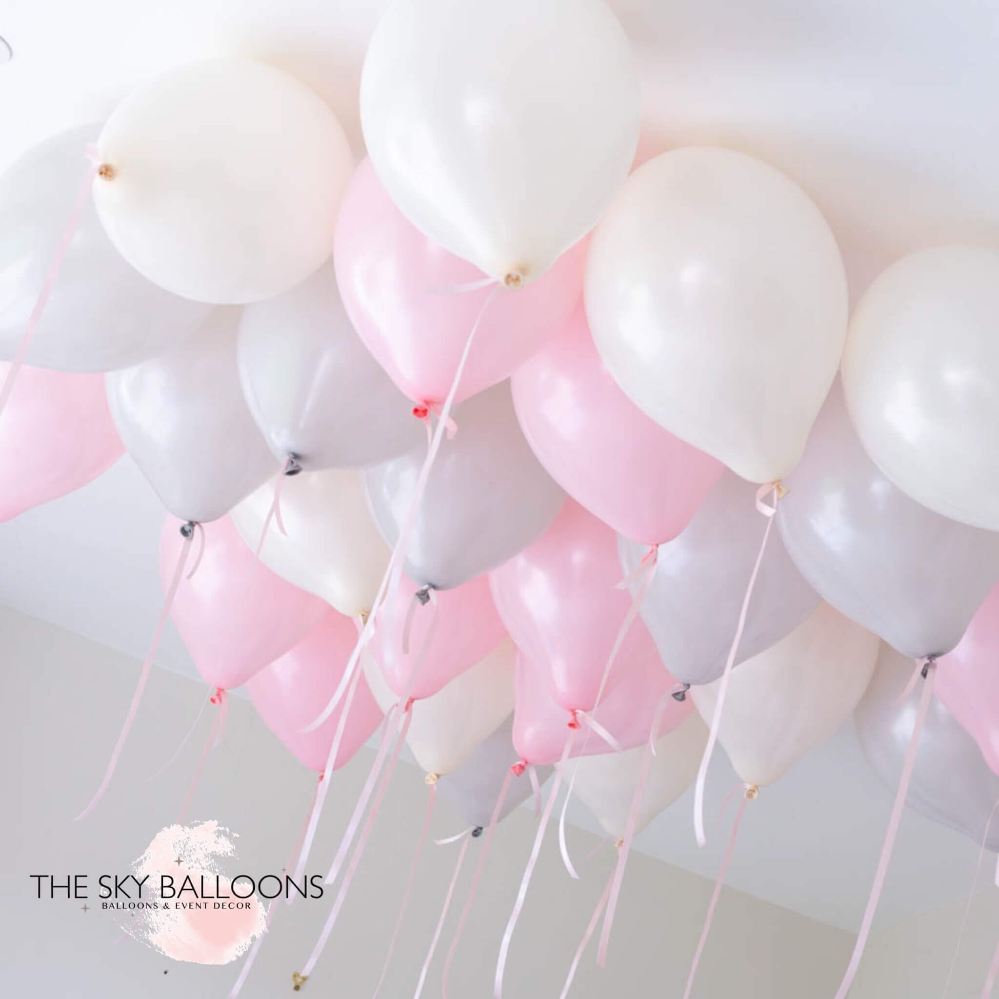 A cluster of helium-filled ceiling balloons in shades of pink, white, and gray, attached to each other and floating against a ceiling.