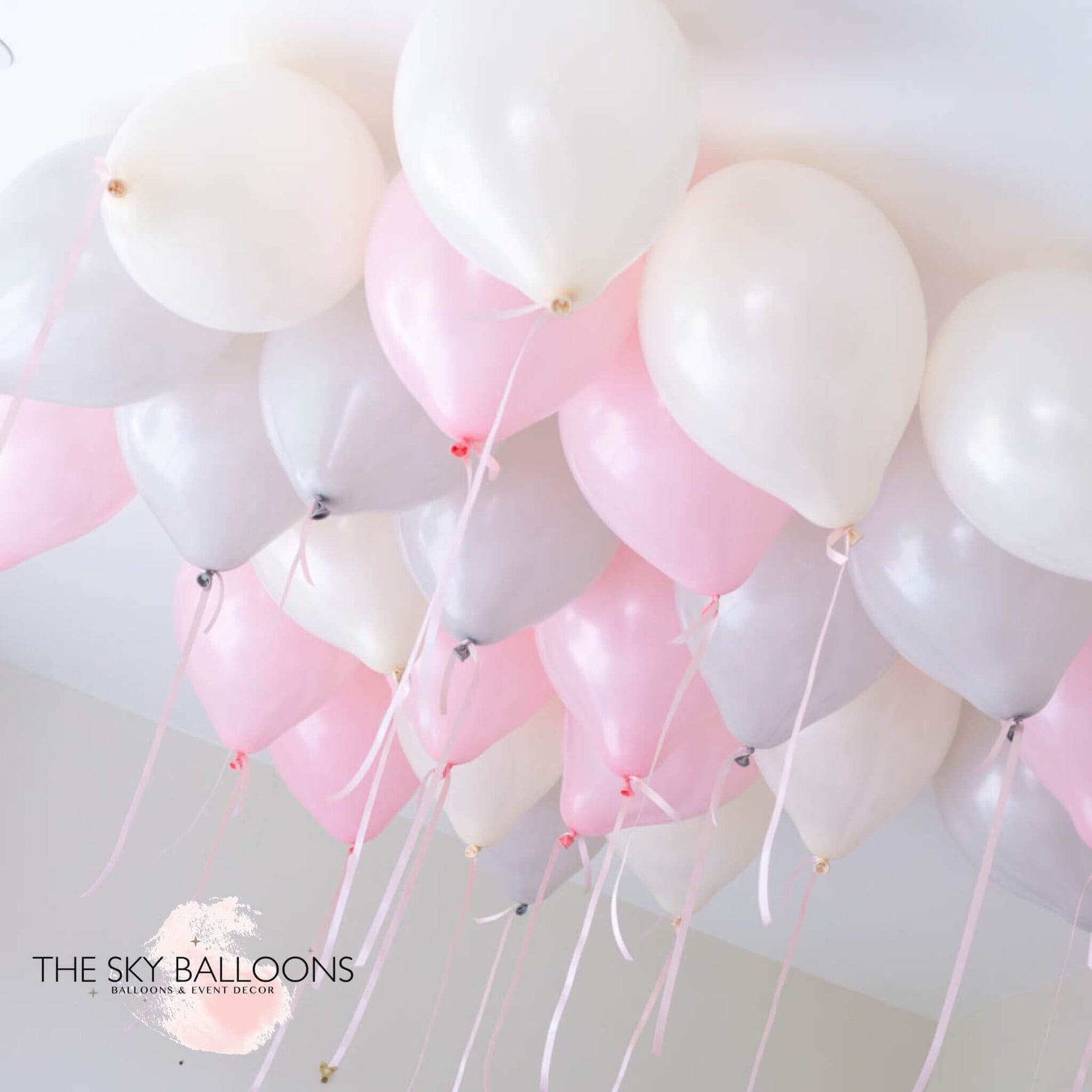 A cluster of helium-filled ceiling balloons in shades of pink, white, and gray, attached to each other and floating against a ceiling.